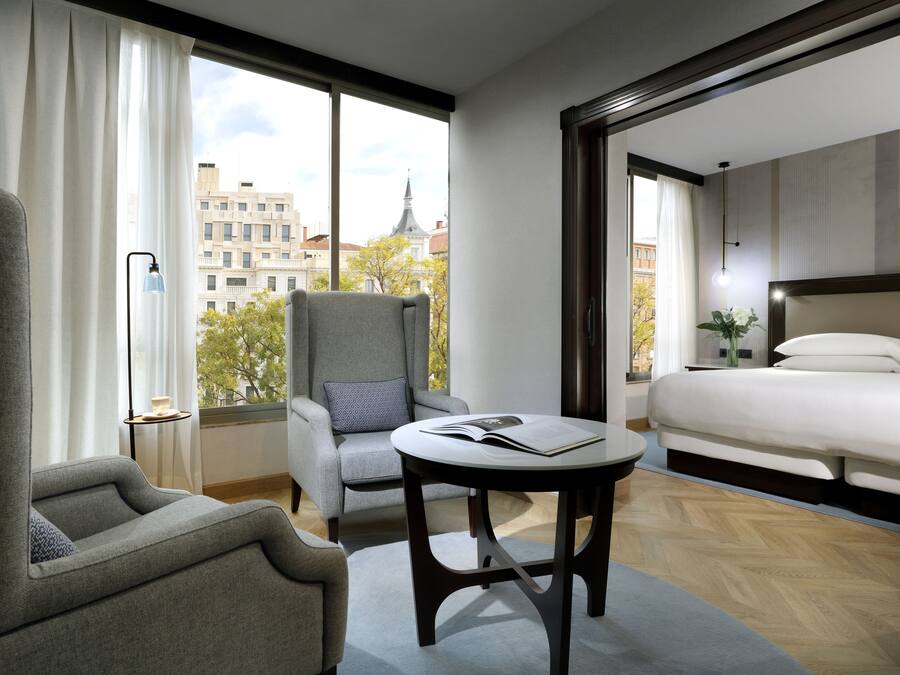 Una habitación de hotel moderna con un gran ventanal, una cama, un área de descanso con una mesa y una silla.