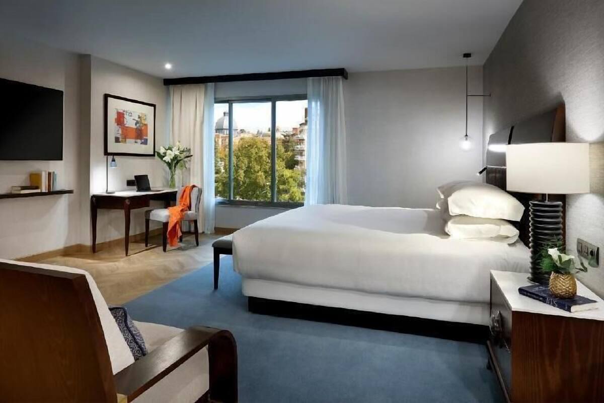 A hotel room with a bed, desk, chair, TV, window, and lamp.