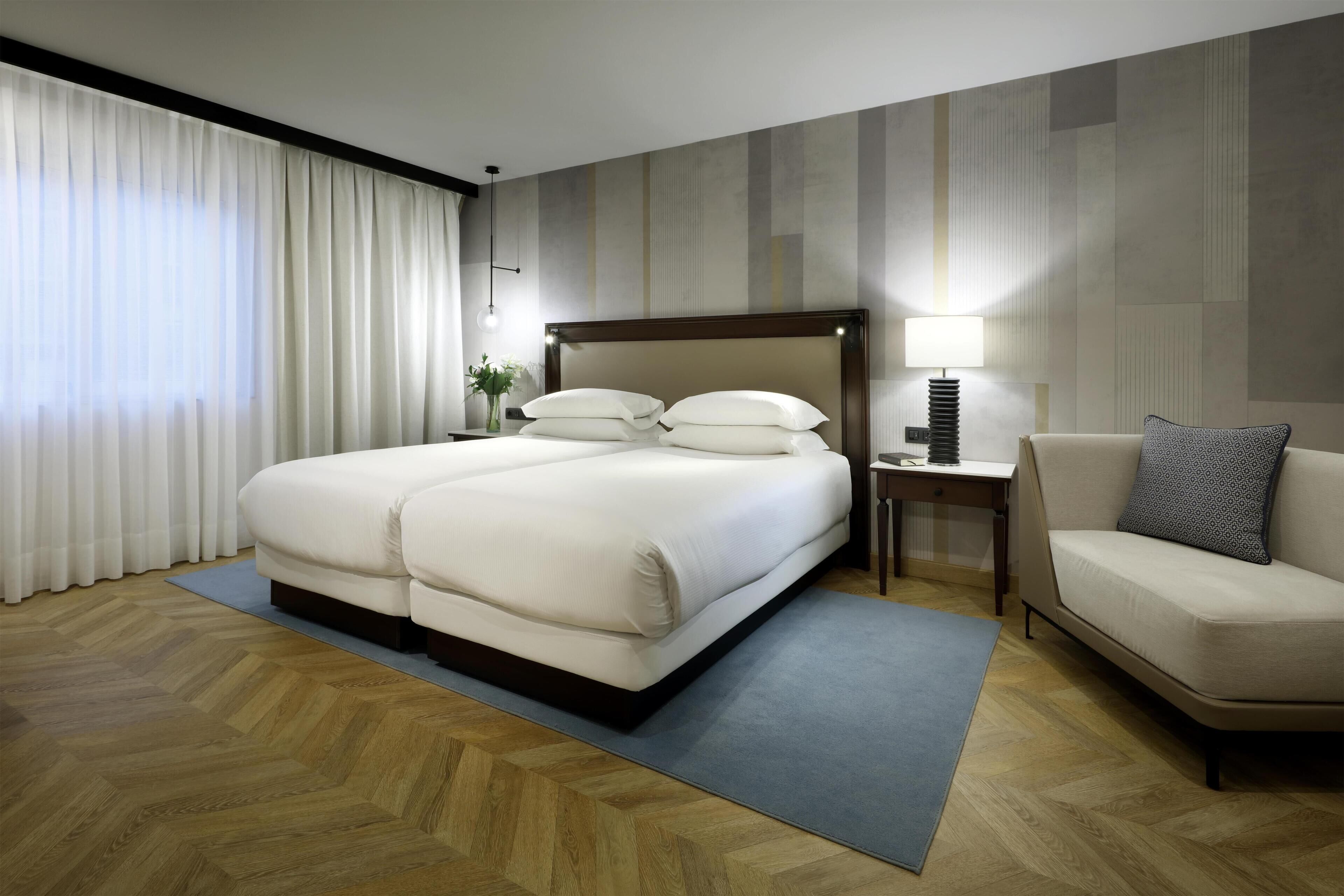 A hotel room with a large bed, a bedside table with a lamp, a sofa, and a view of the outdoors through sheer curtains.