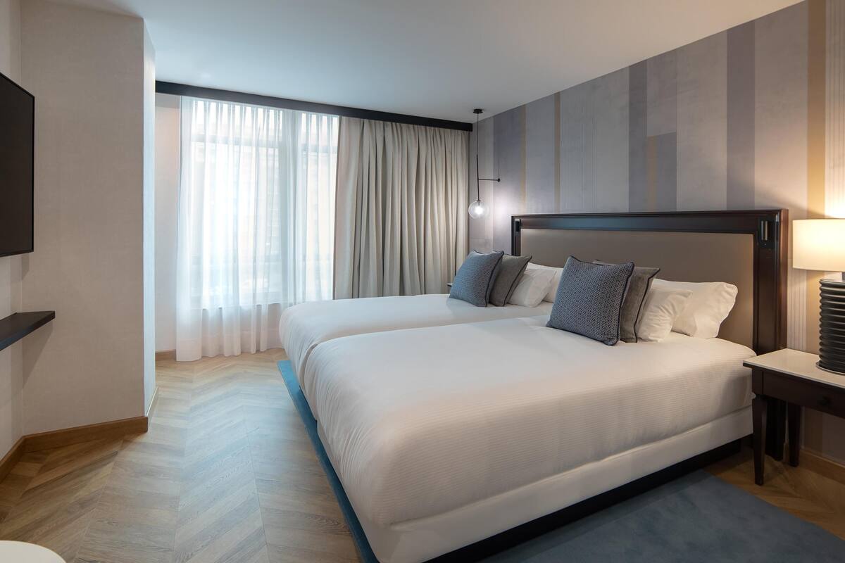 A hotel room with two beds, a TV, a lamp, and a large window with curtains.