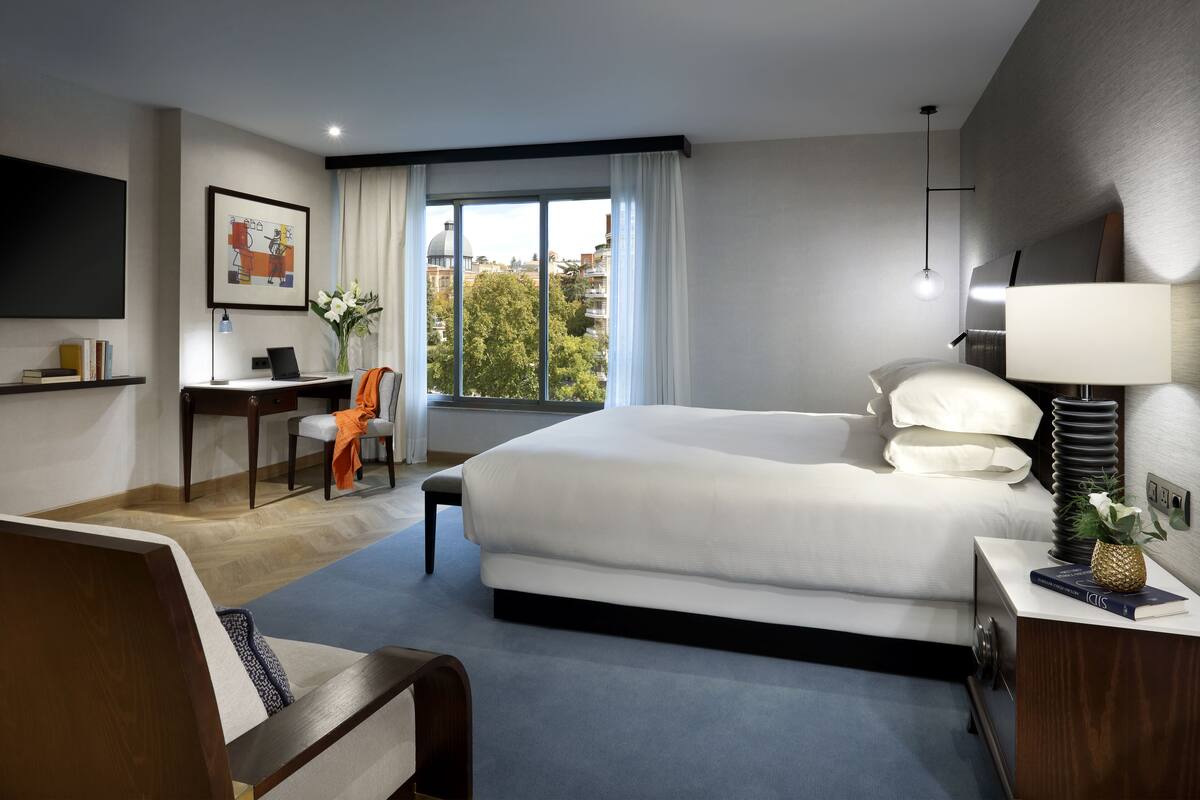A modern hotel room with a large bed, a desk with a chair, a TV, and a window with curtains.