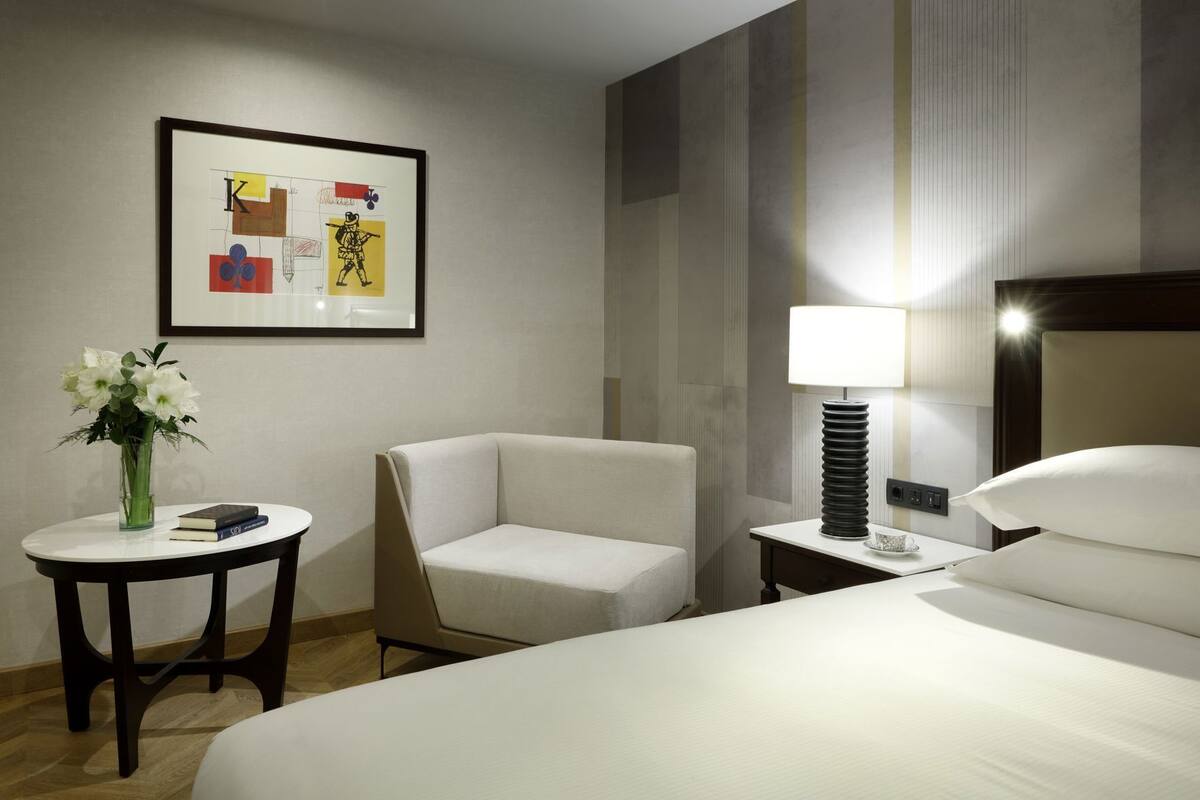 A hotel room with a bed, a chair, a small table with a vase and books, a lamp, and a framed picture on the wall.