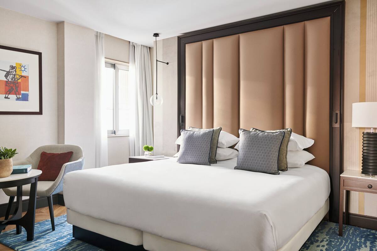 A hotel room with a large bed, a chair, a small table with a plant, a lamp, and a window with curtains.
