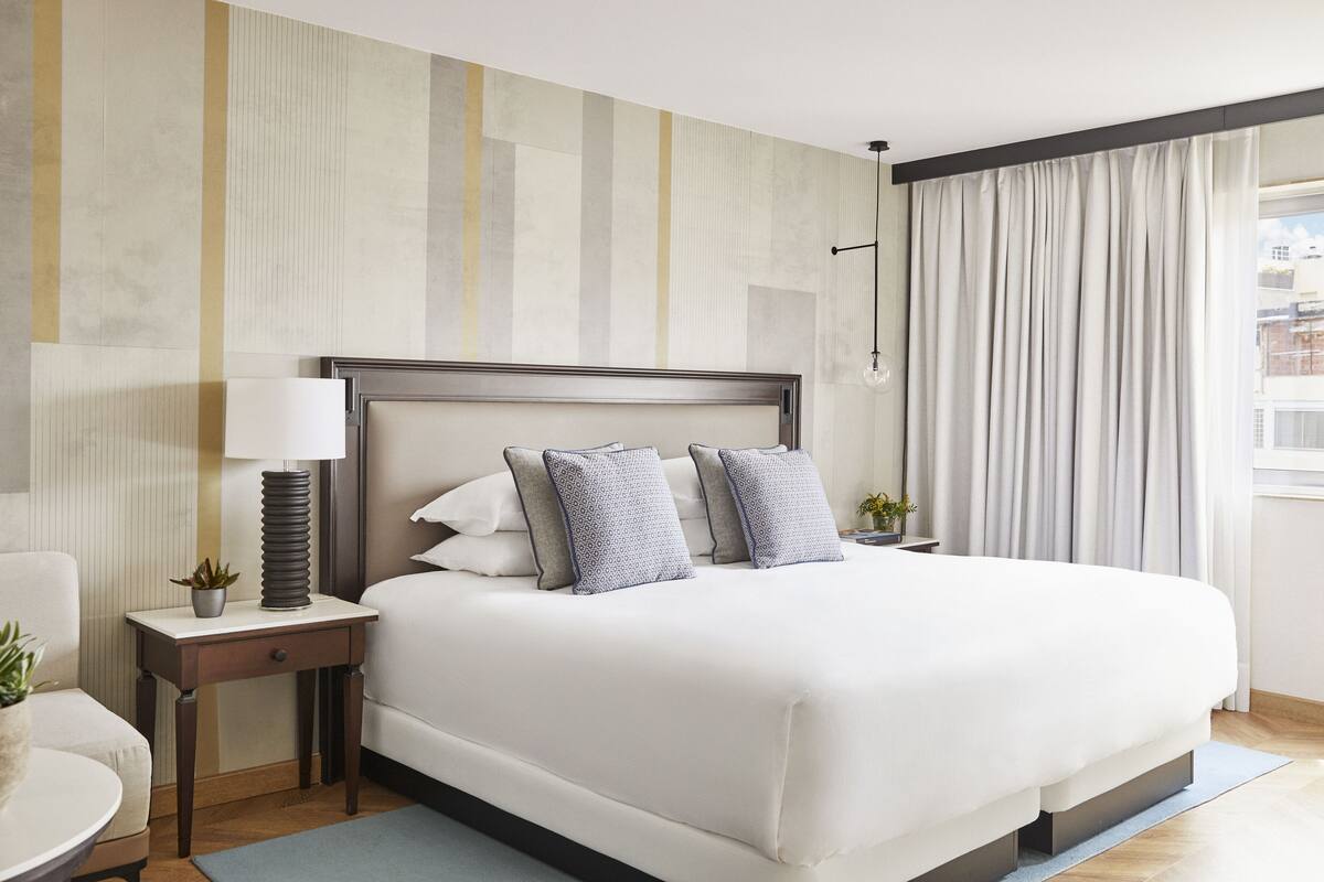 A modern hotel room with a large bed, a bedside table with a lamp, a small plant, and a view of the city through the window.