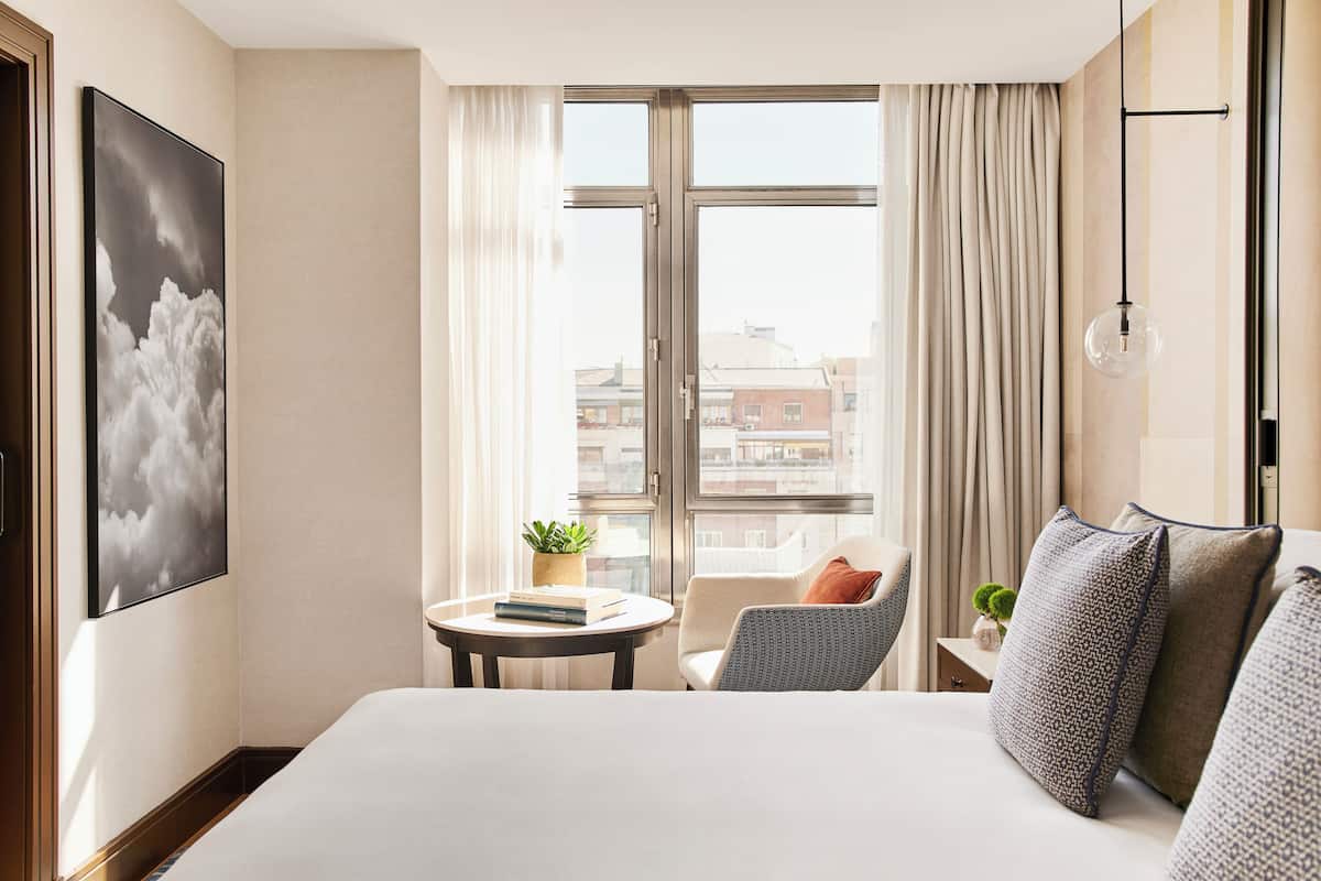 A hotel room with a large bed, a small round table with a plant, a chair, and a view of buildings through a large window with curtains.