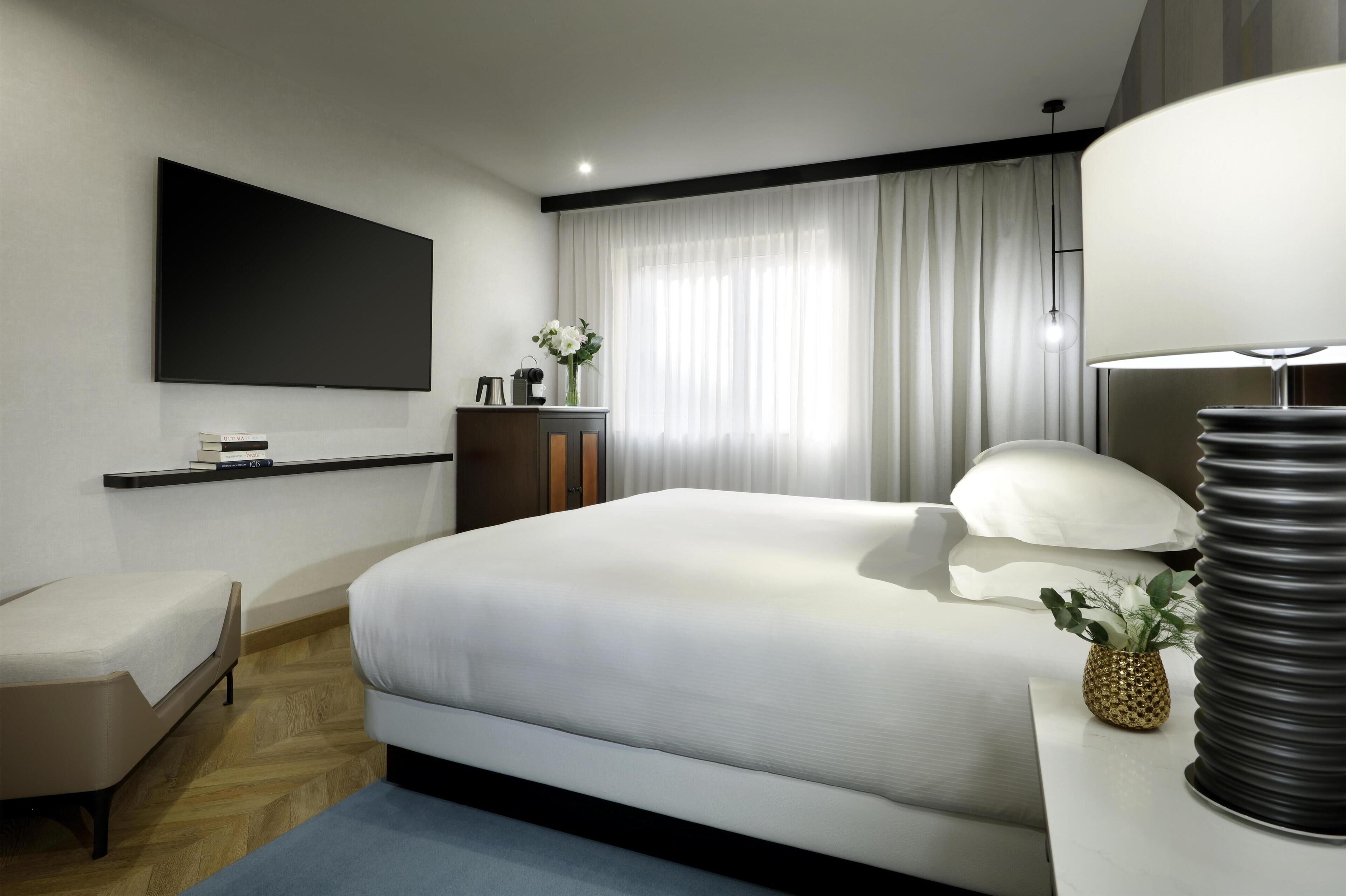 Moderni hotellihuone, jossa on suuri sänky, seinälle asennettu taulutelevisio, pieni työpöytä lampun kanssa ja ikkuna, jossa on ohuet verhot.