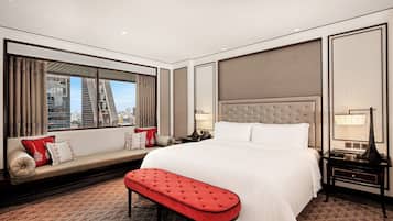 A hotel room with a large bed, a bench, a nightstand, a lamp, and a window with a city view.