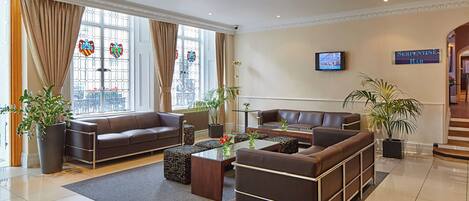 A modern hotel lobby with leather sofas, a coffee table, potted plants, and a television on the wall.