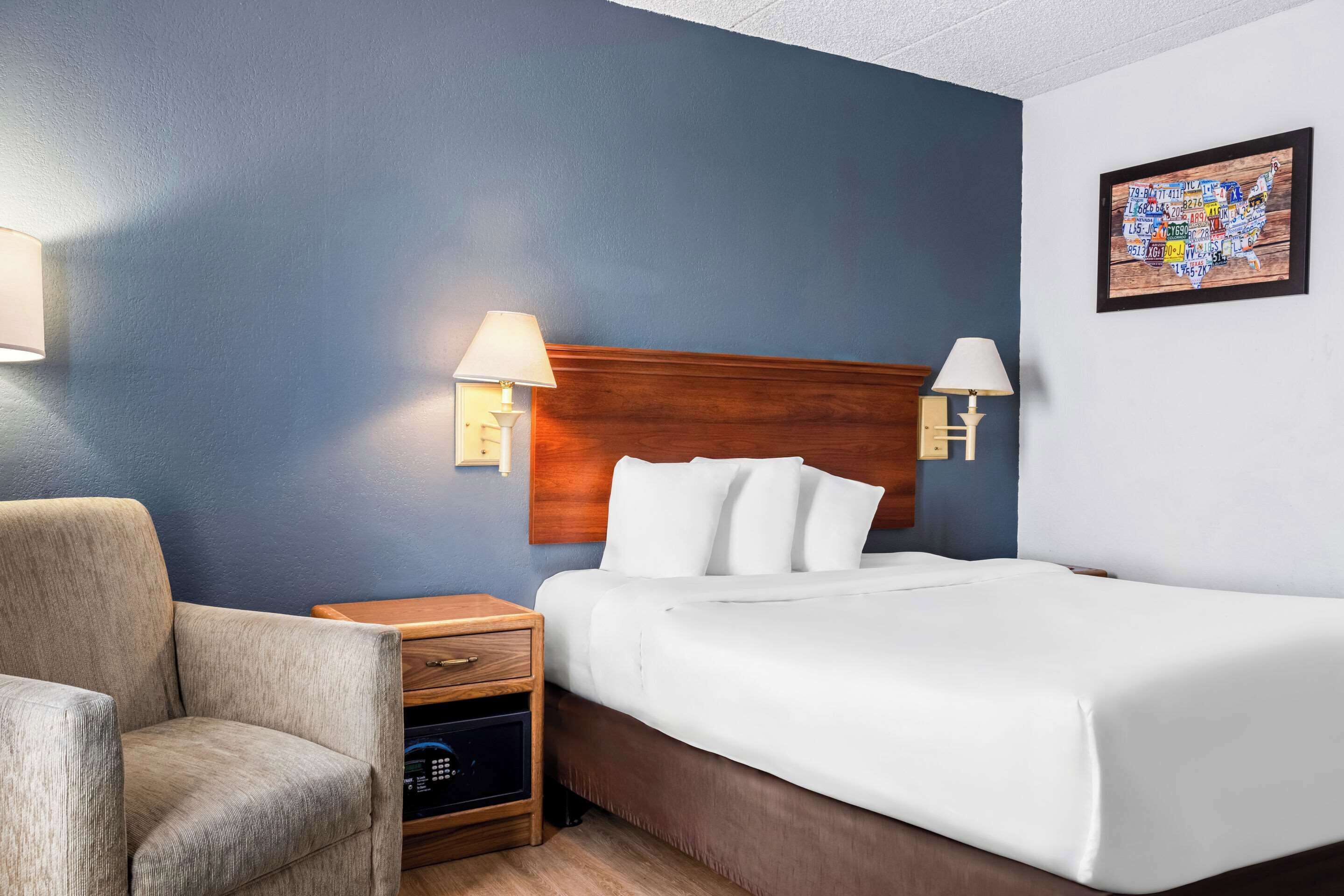 A hotel room with a bed, two bedside lamps, a nightstand, a chair, and a wall art piece.