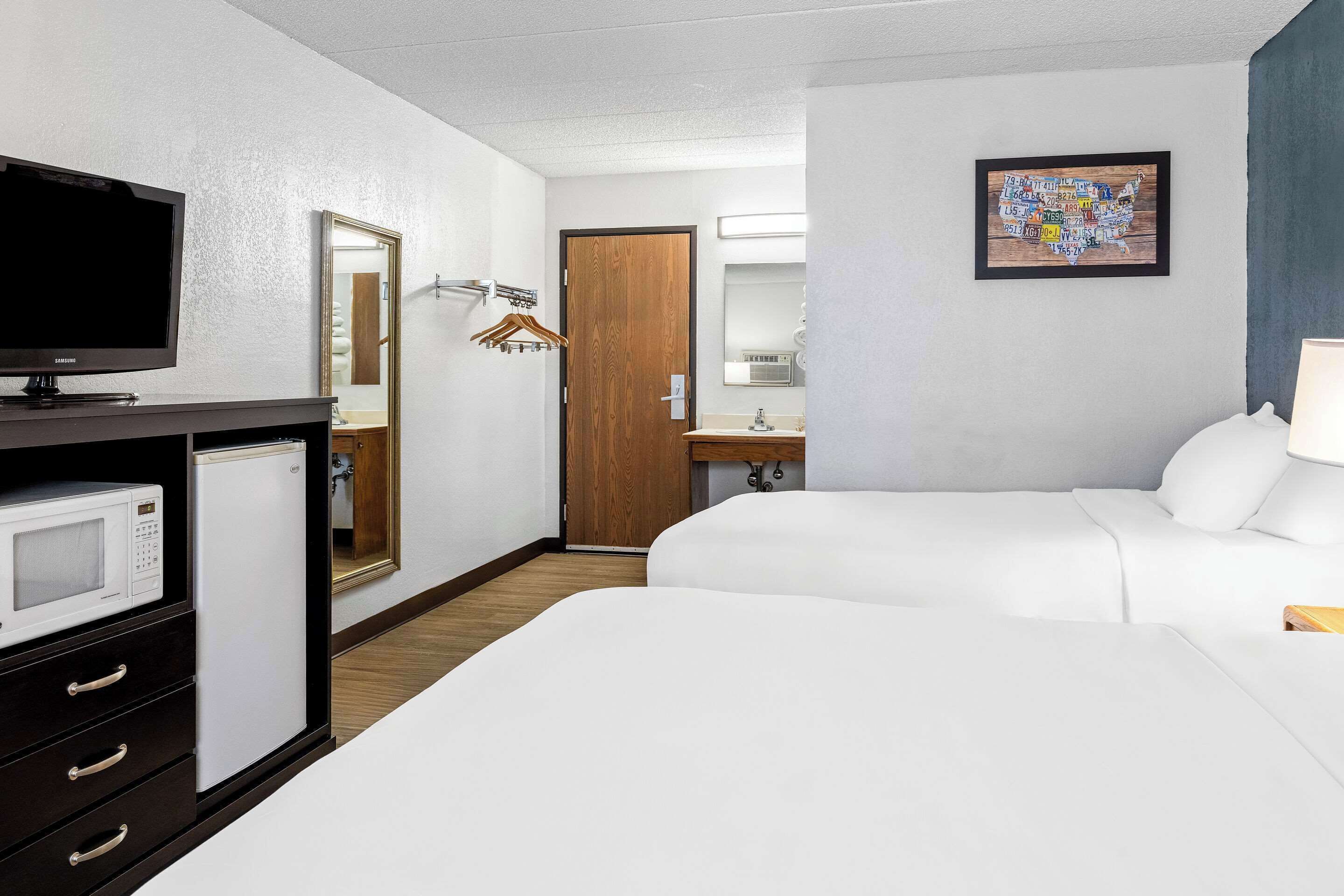 Habitación de hotel con dos camas, un televisor, un microondas, un aparador, una puerta de madera, un baño con espejo y una obra de arte con un mapa en la pared.