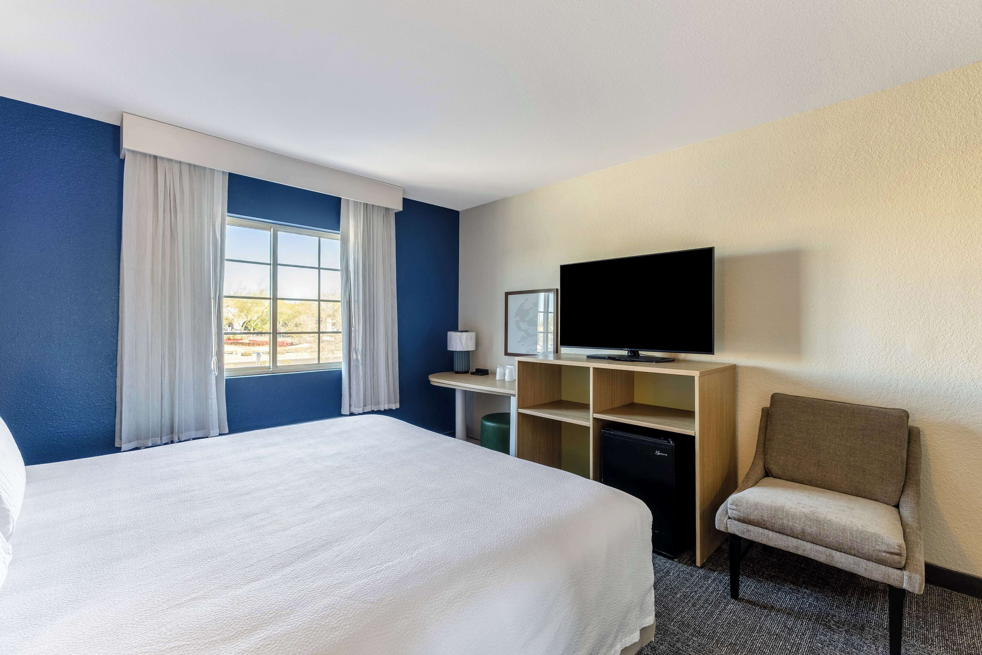 A hotel room with a large bed, a television, a desk with a lamp, and a chair.