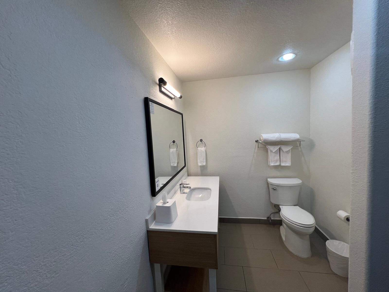 Un baño pequeño y limpio con un lavamanos, espejo e inodoro blancos. Hay un perchero sobre el inodoro y un cubo de basura al lado.