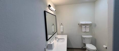 Un baño pequeño y limpio con un lavamanos, espejo e inodoro blancos. Hay un perchero sobre el inodoro y un cubo de basura al lado.