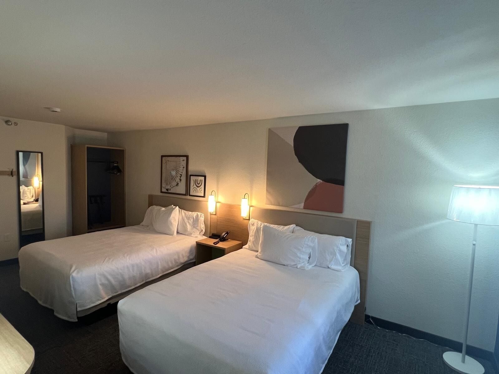 A hotel room with two beds, a mirror, a nightstand with a phone, and a wall-mounted light fixture.
