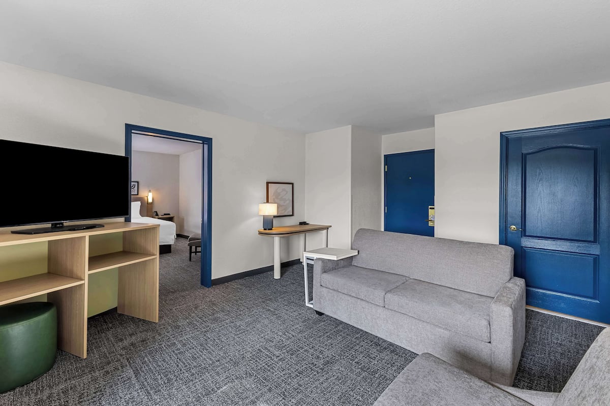 A hotel room with a flat-screen TV, a grey sofa, a small white table, a lamp, and a blue door.
