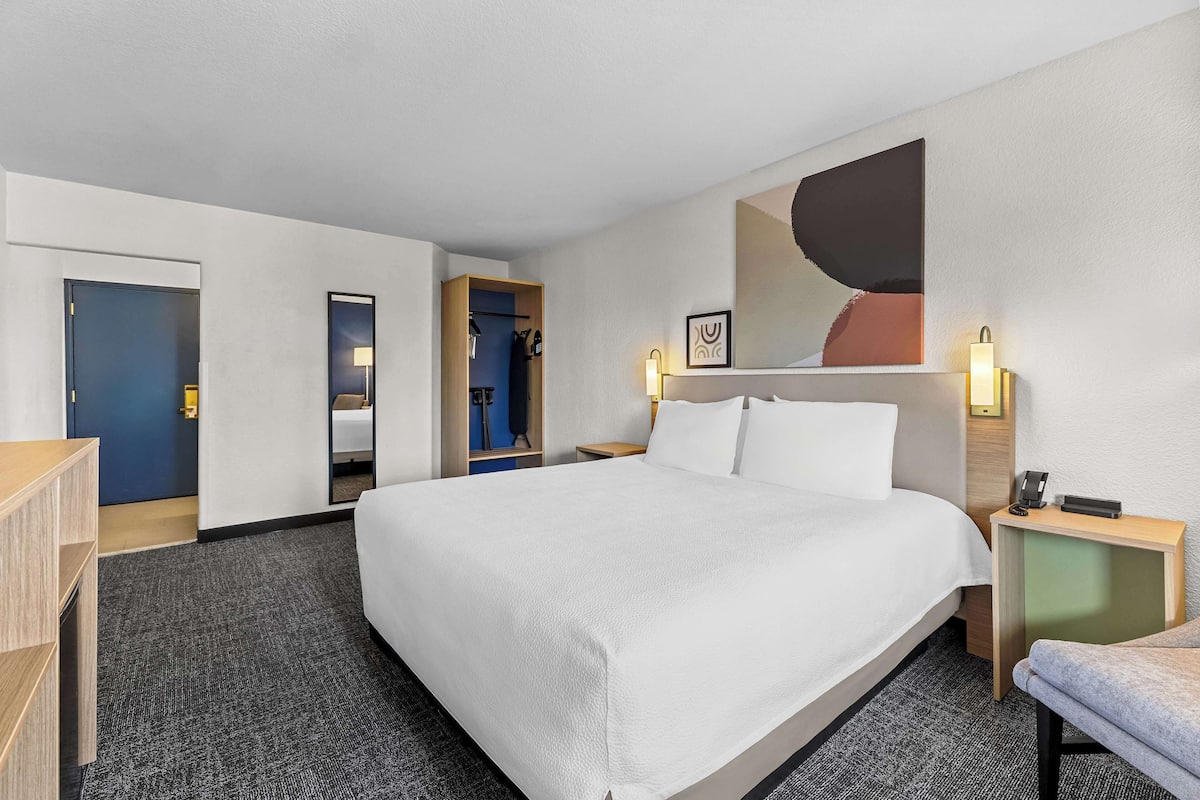 A hotel room with a large bed, a desk, a chair, a wardrobe, and a mirror.