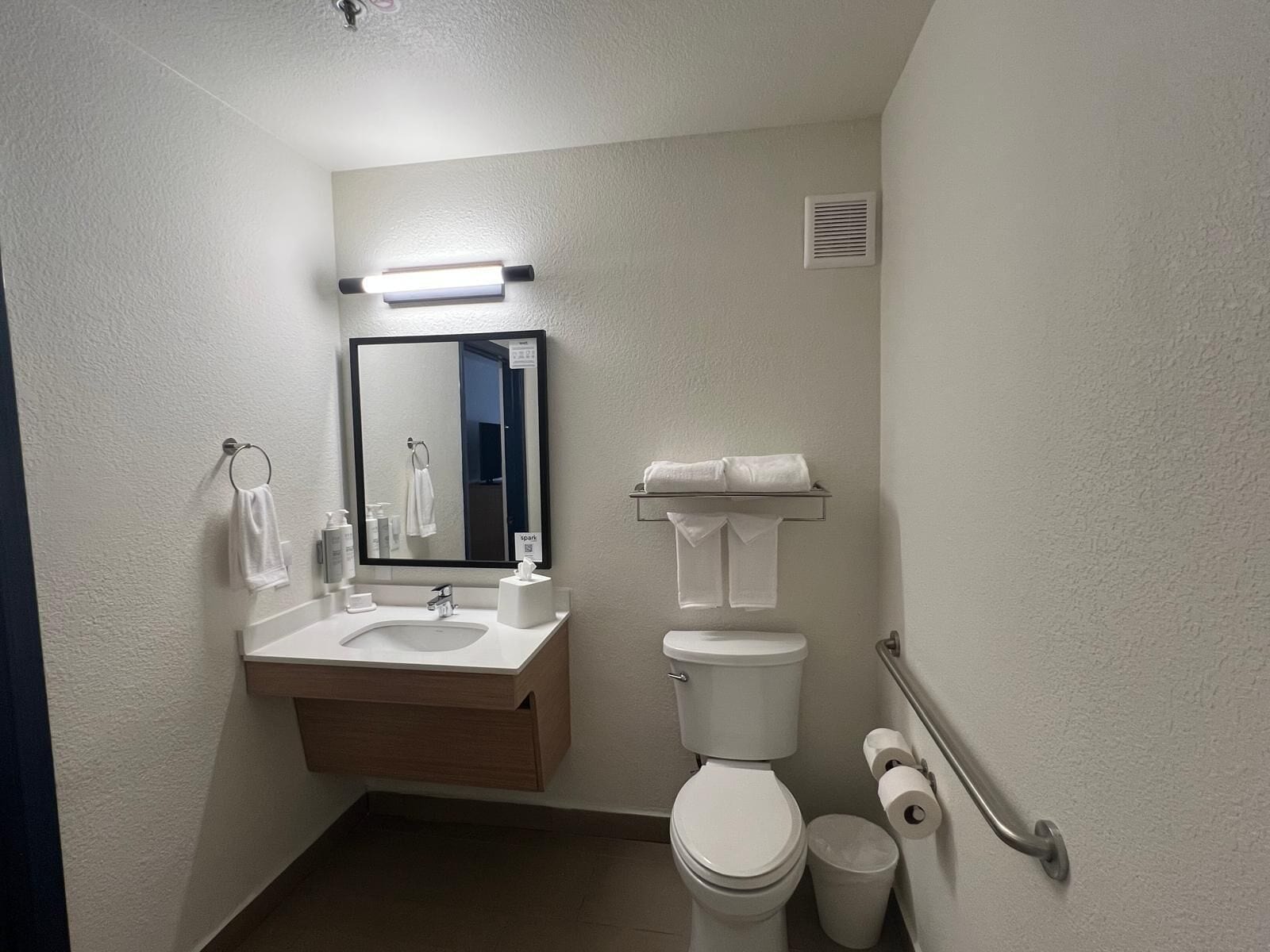 A compact hotel bathroom with a sink, mirror, toilet, and handrail.