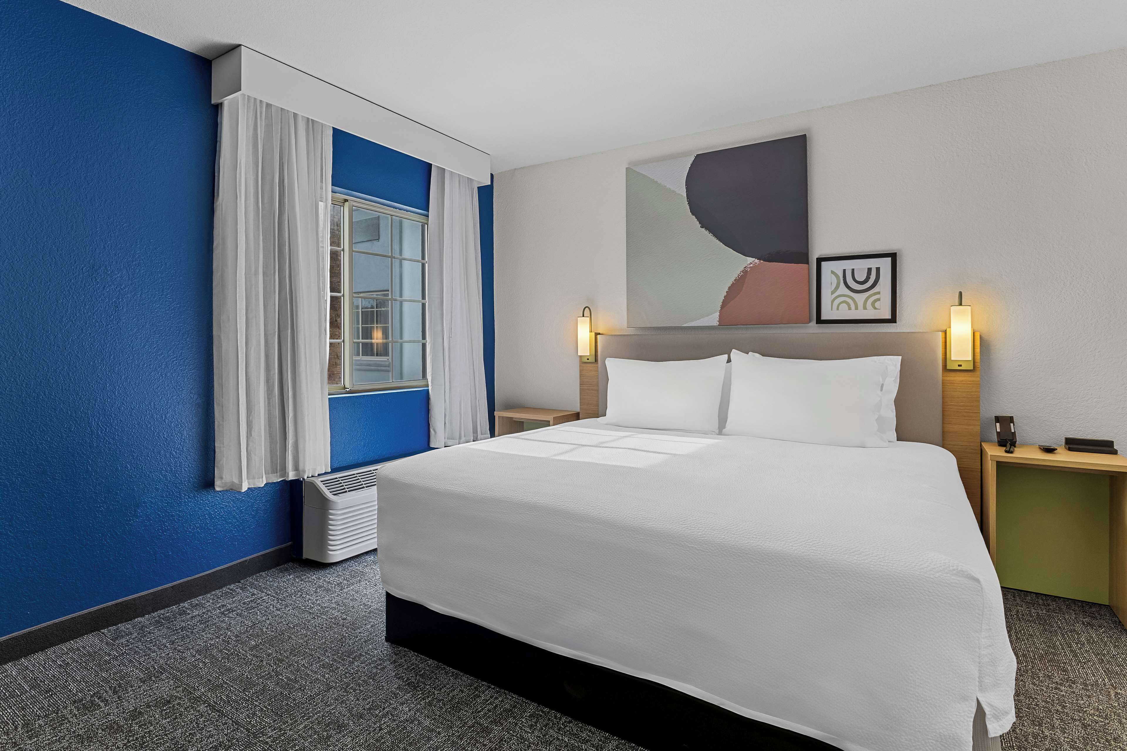 A hotel room with a large bed, a window with white curtains, a wall-mounted artwork, and a bedside table with a lamp.