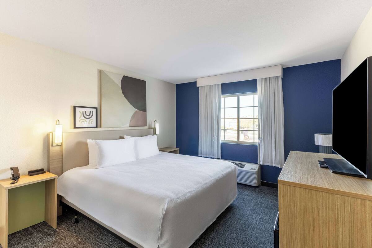 A hotel room with a large bed, a desk, a flat-screen TV, a window with curtains, and a wall-mounted artwork.