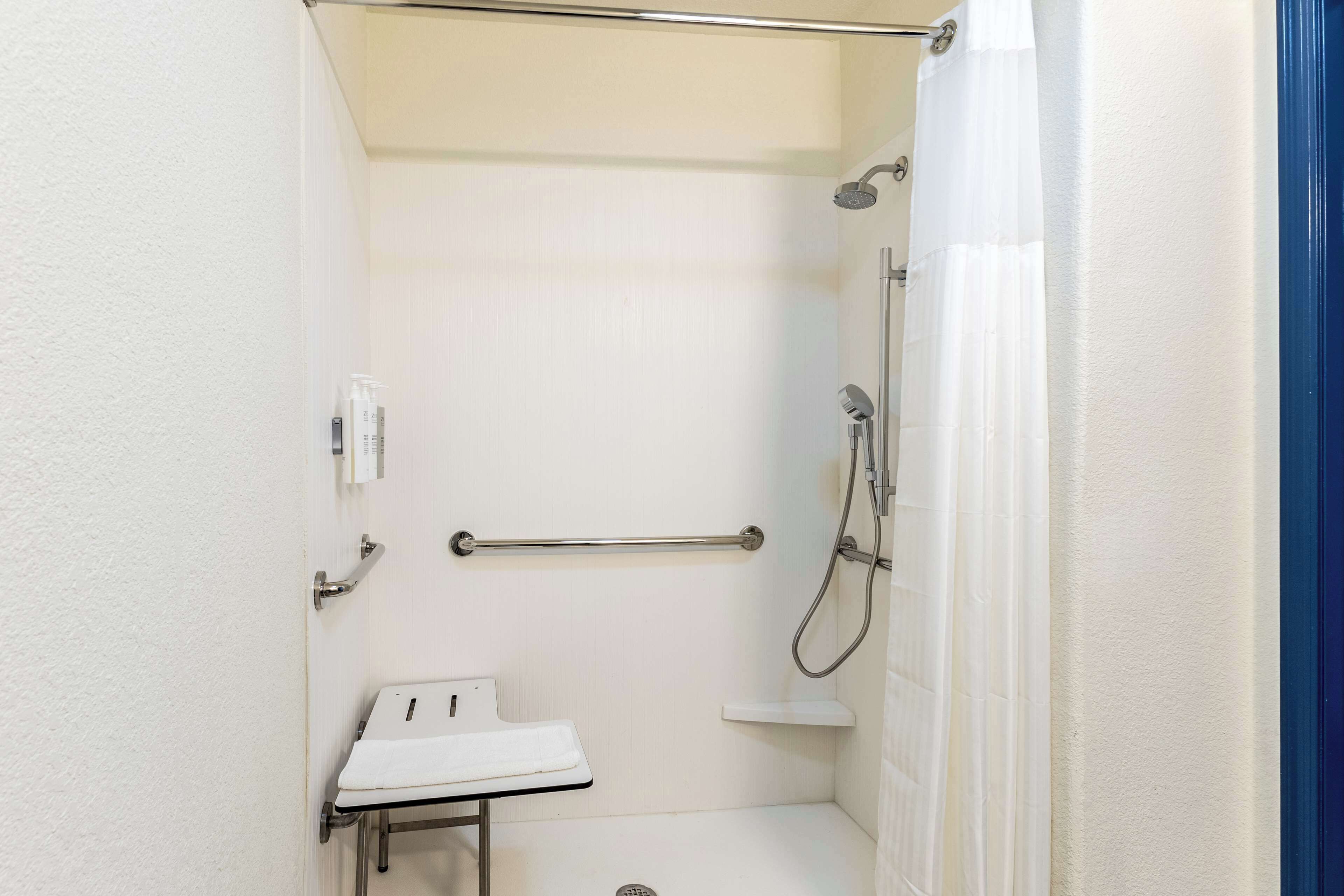 A small, white, walk-in shower with a grab bar, a white sink, and a showerhead.