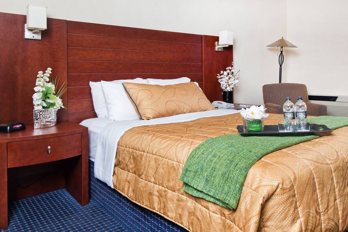 Habitación de hotel con cabecera de madera, cama con colcha estampada, mesita de noche con control remoto, lámpara y silla.