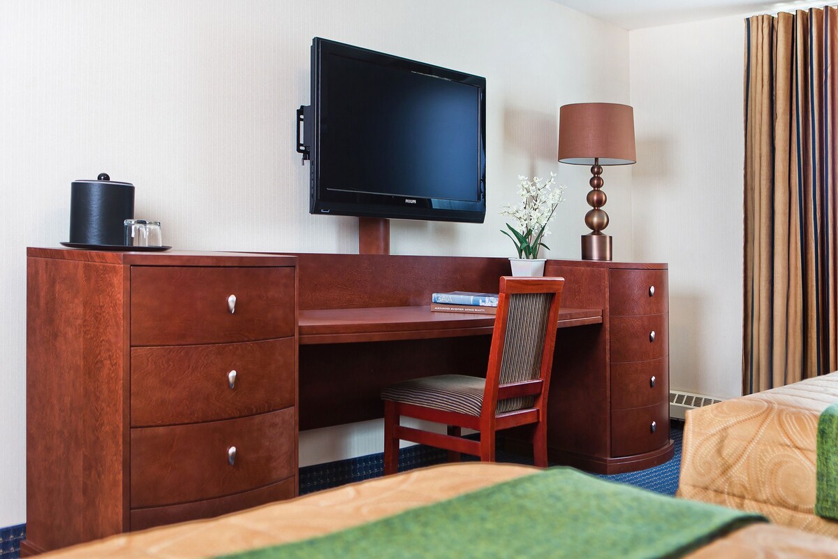 Habitación de hotel con escritorio de madera, televisor, lámpara, silla, cama con manta verde y ventana con cortinas.