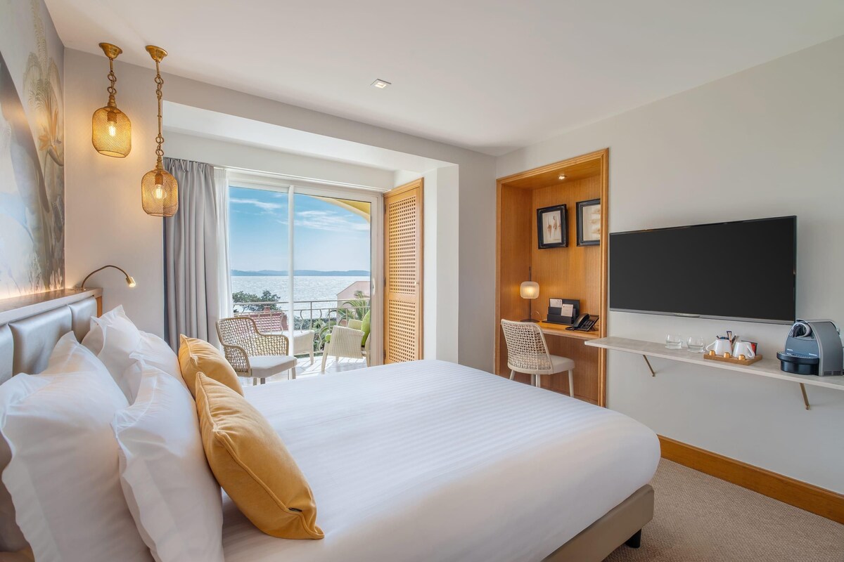 A hotel room with a large bed, a desk with a chair, a TV, and a balcony with a view.
