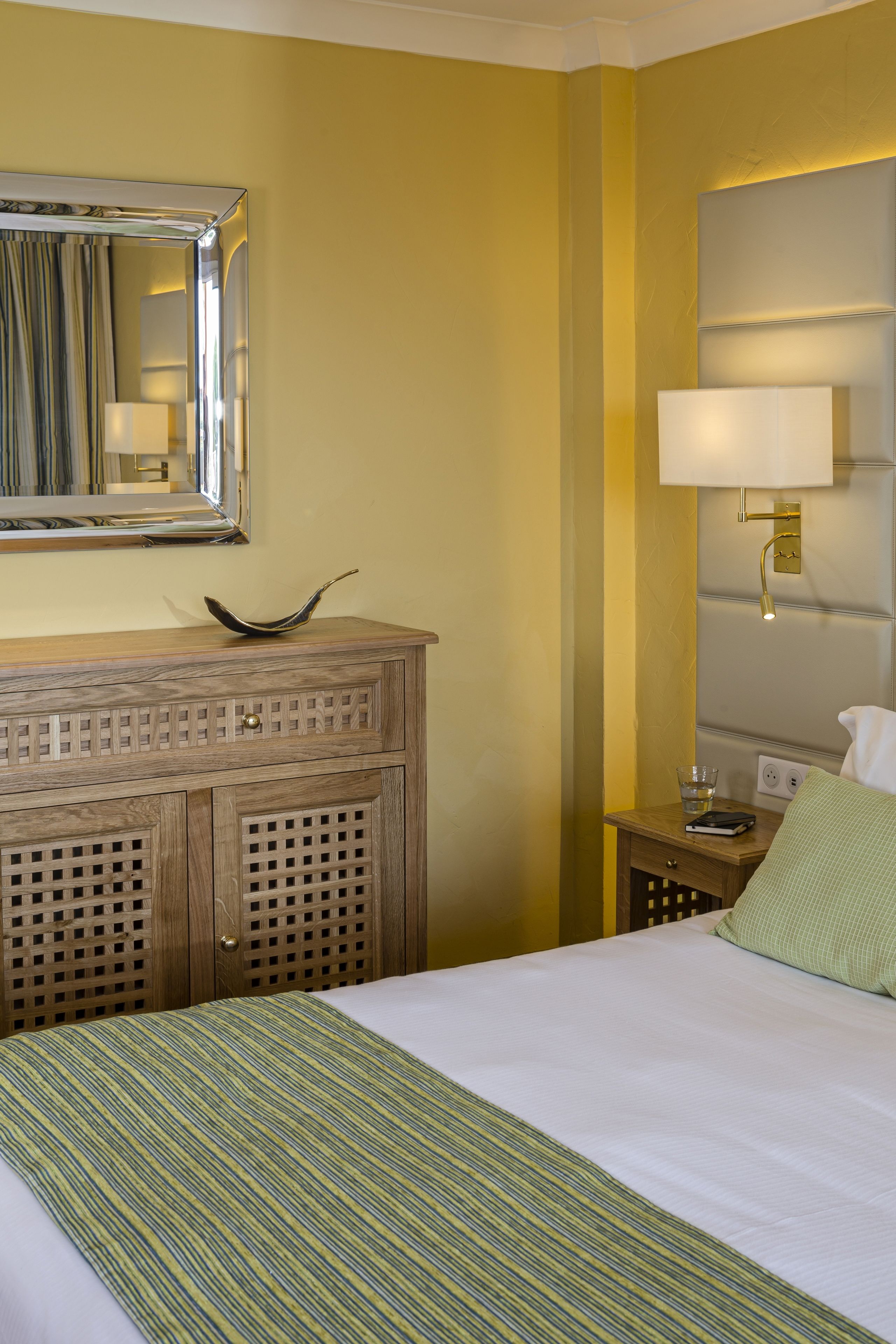 A hotel room with a bed, a wooden dresser, a mirror, a bedside lamp, and a green pillow.