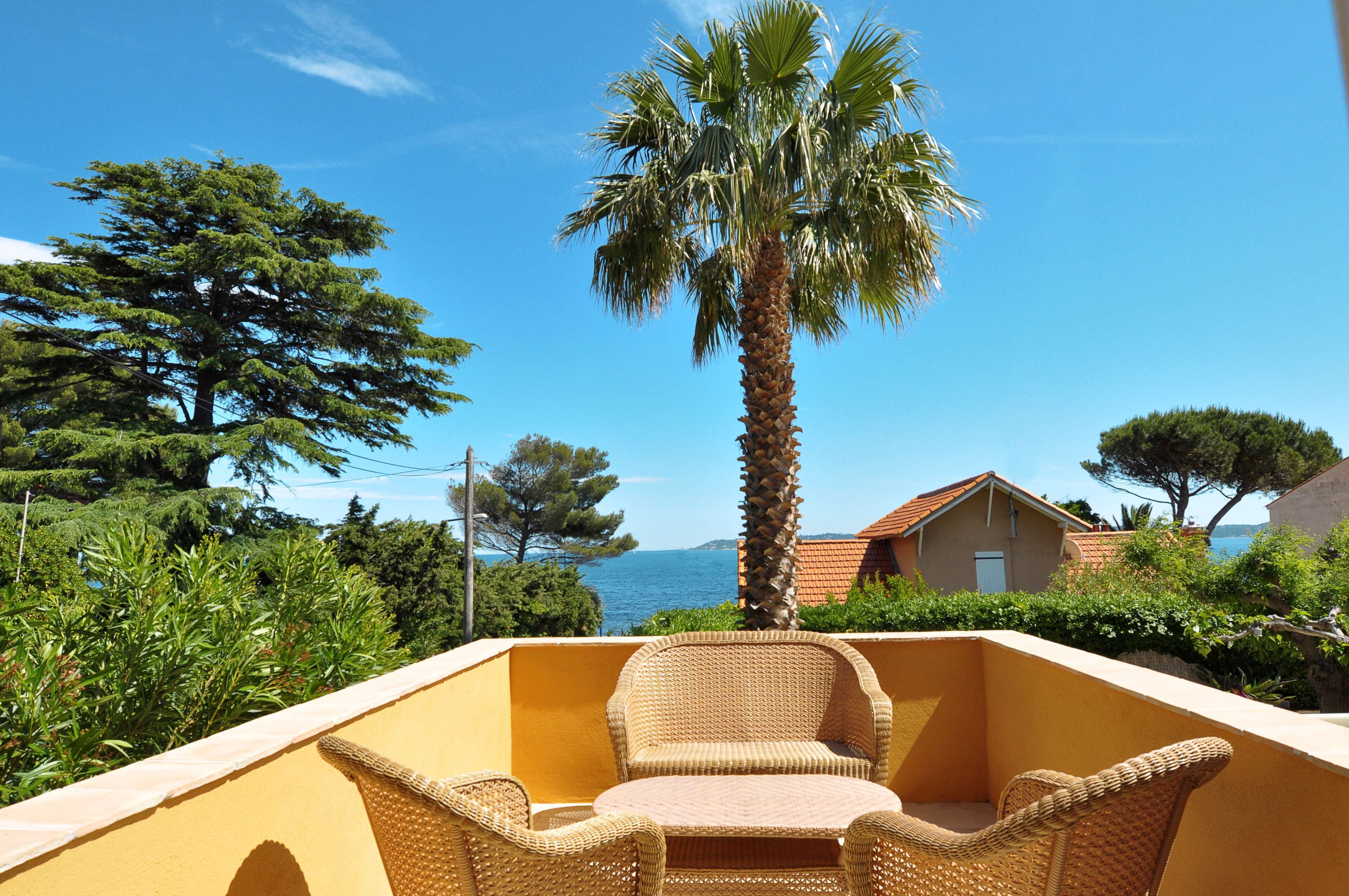 A balcony with wicker furniture, a palm tree, and a view of the sea and houses.