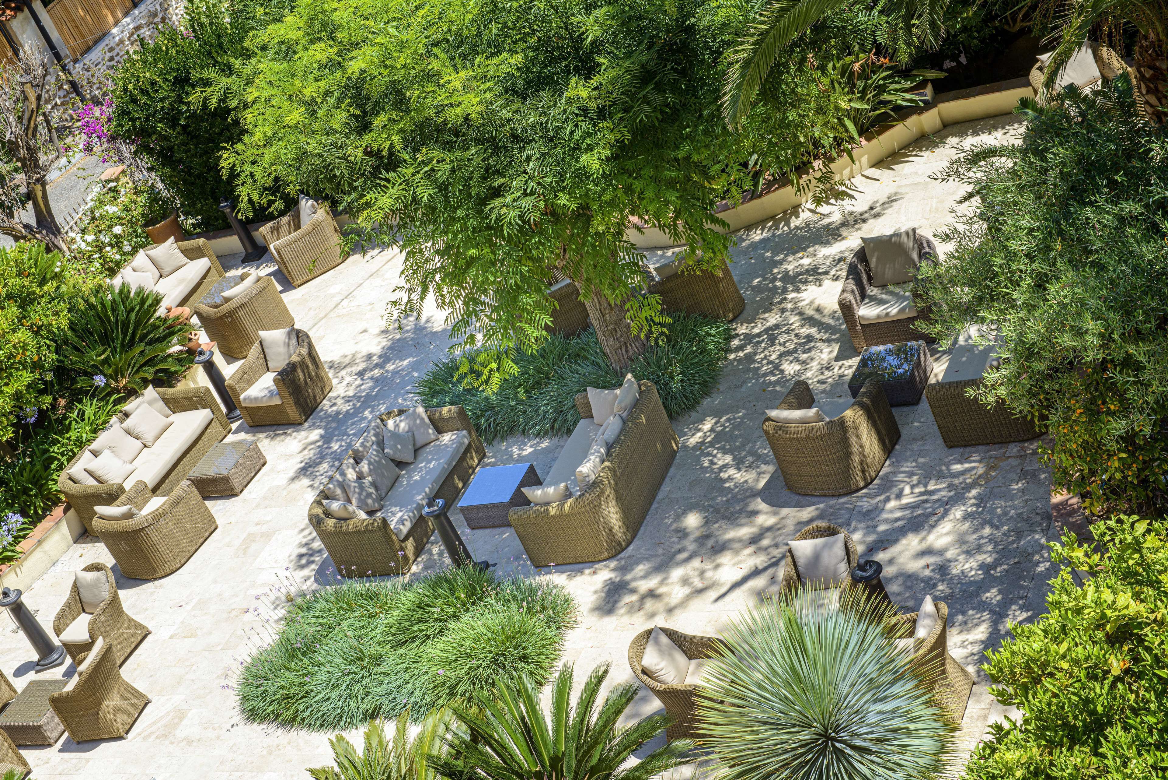 An outdoor patio area with wicker furniture, including sofas and chairs, surrounded by various plants and trees.