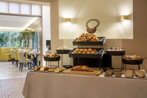 A buffet with various breads and pastries, a dining area with yellow chairs, and a large window with a view of greenery. - Best Western Premier Montfleuri (Sainte-Maxime)