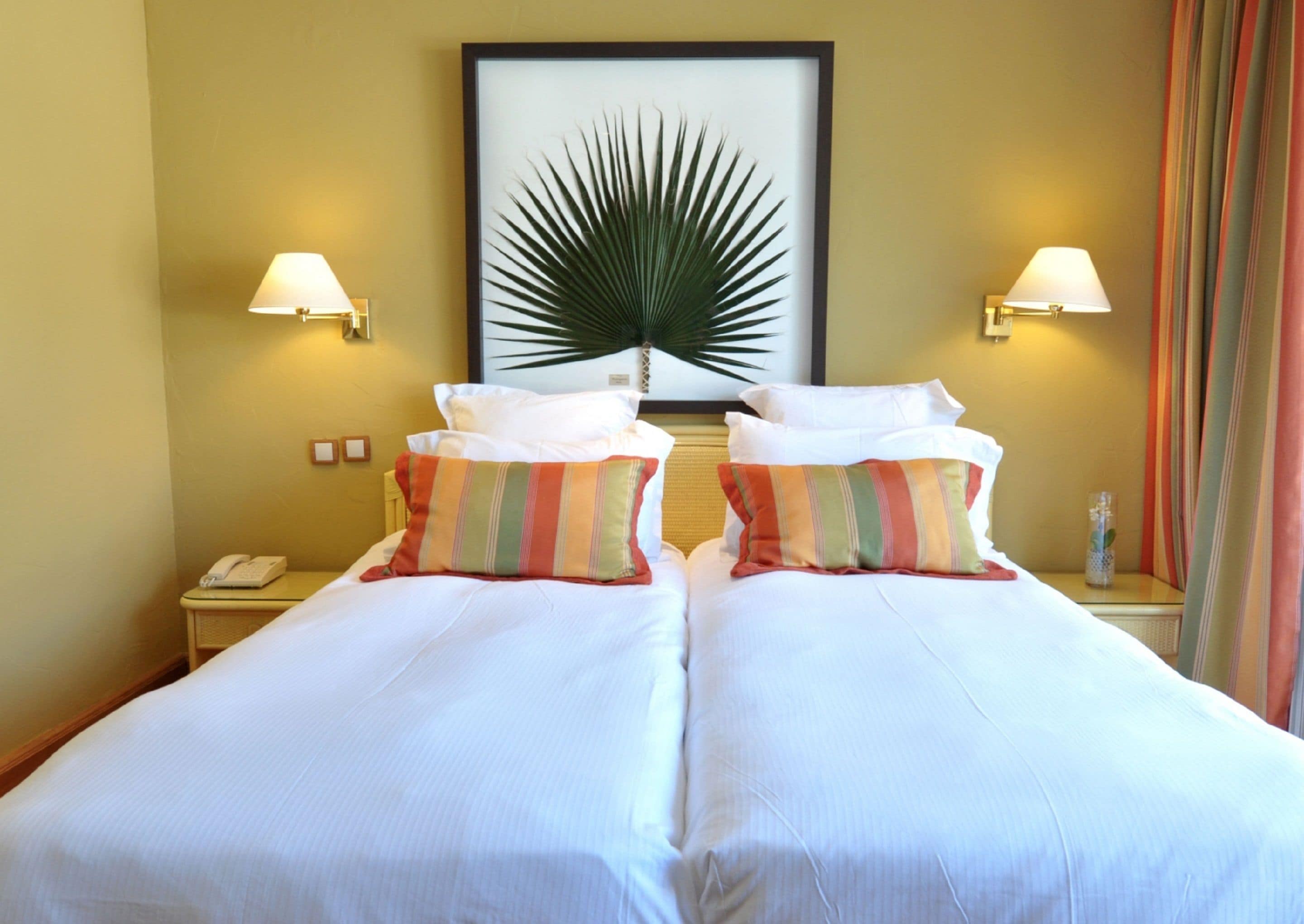 A double bed with white linens and striped pillows, a framed palm leaf artwork on the wall, and two bedside lamps.