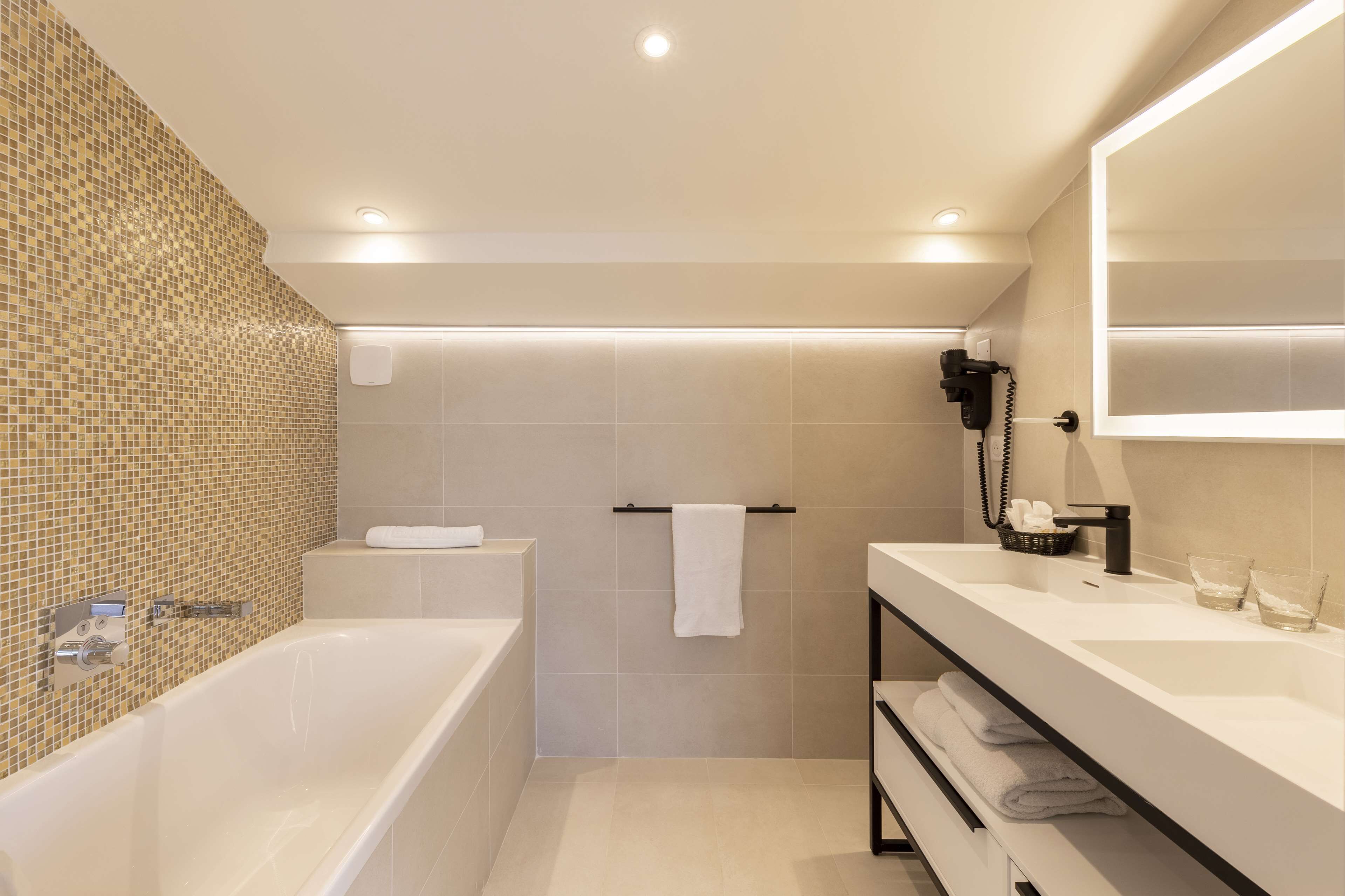 Une salle de bain moderne avec une baignoire, un meuble-lavabo avec un miroir et un sèche-cheveux fixé au mur.