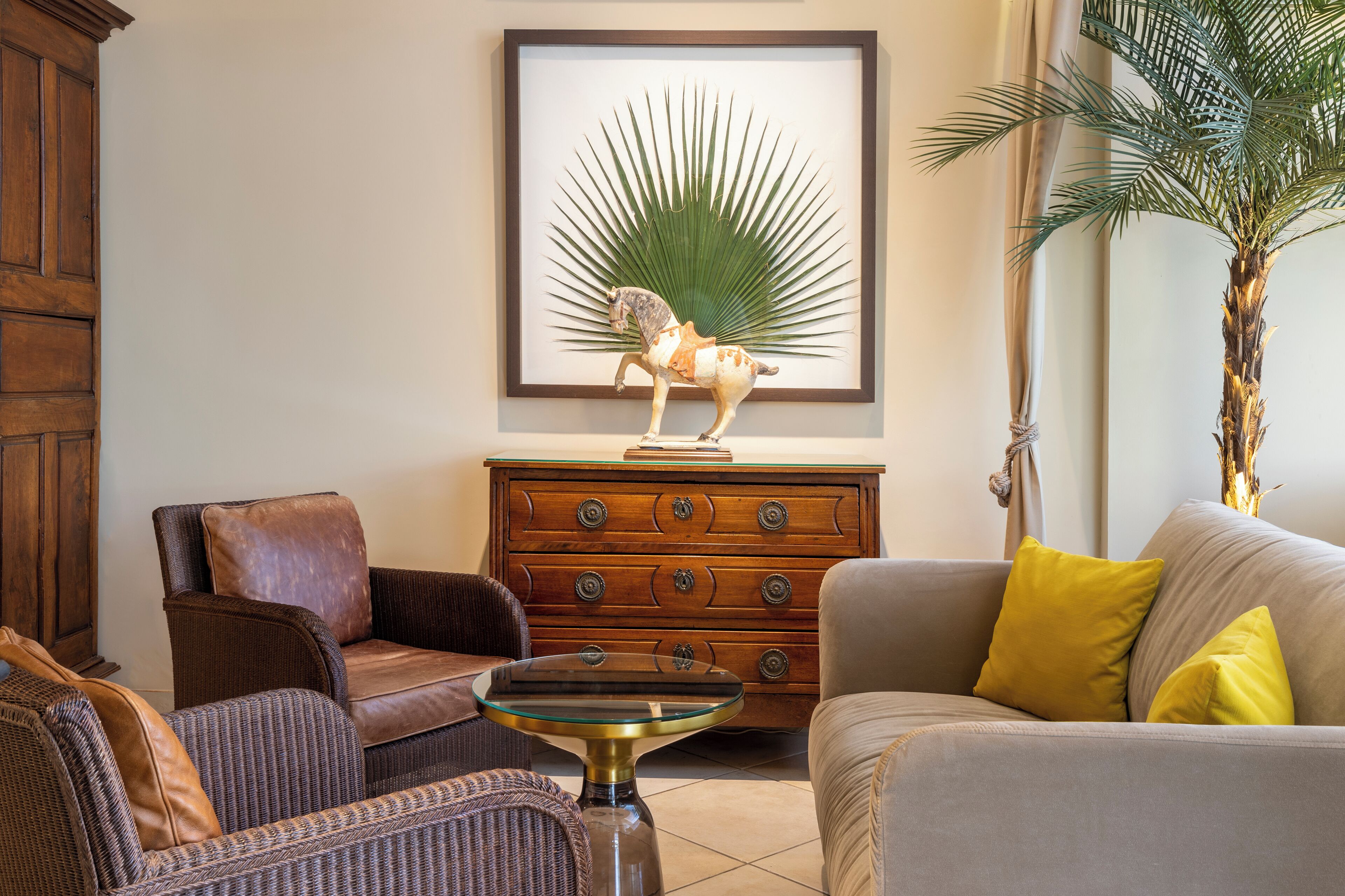 A living room with a wooden cabinet, a framed artwork of a horse, a sofa with yellow cushions, and a potted palm tree.