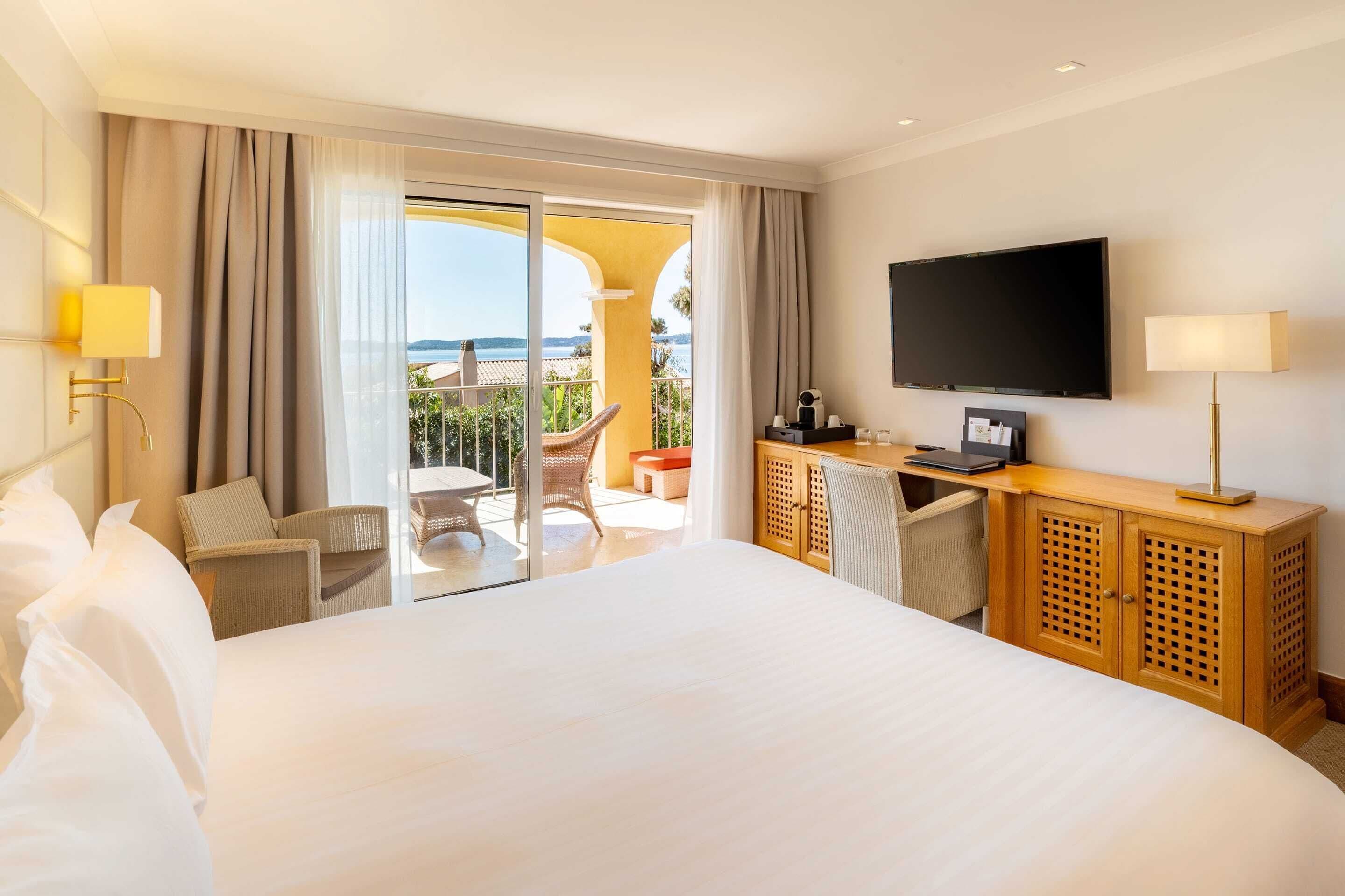 A hotel room with a large bed, a desk with a flat-screen TV, a chair, and a view of the ocean.