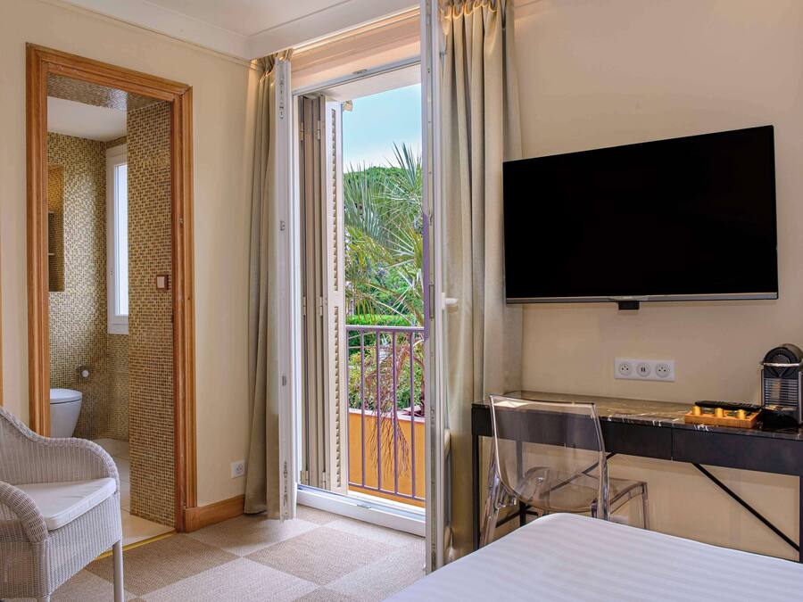 Hotelkamer met een bed, een televisie, een bureau met koffiezetapparaat, een stoel en een schuifdeur naar een balkon met planten.