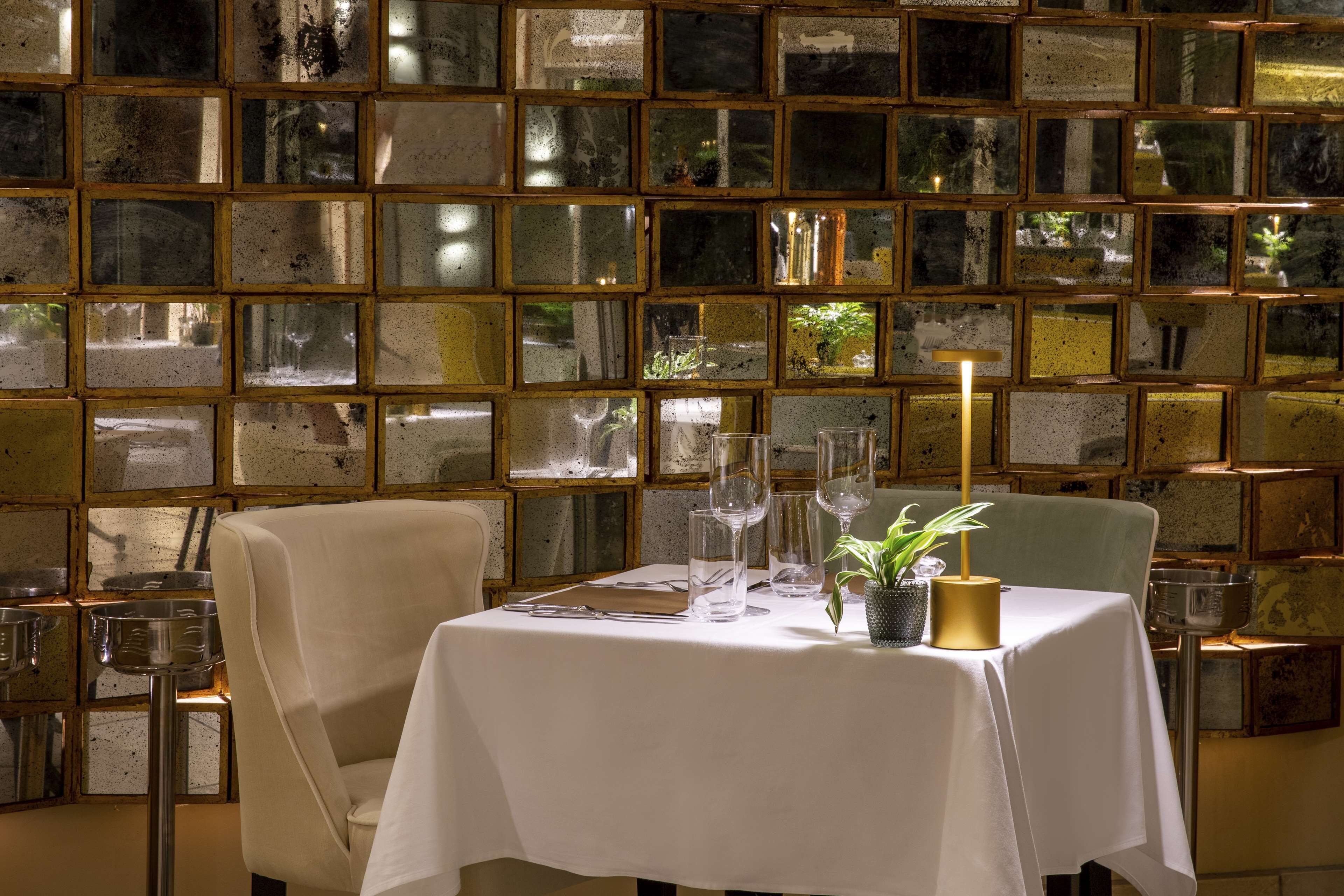 Una mesa de comedor con mantel blanco, rodeada de sillas, y un muro de paneles de vidrio reflectante al fondo.