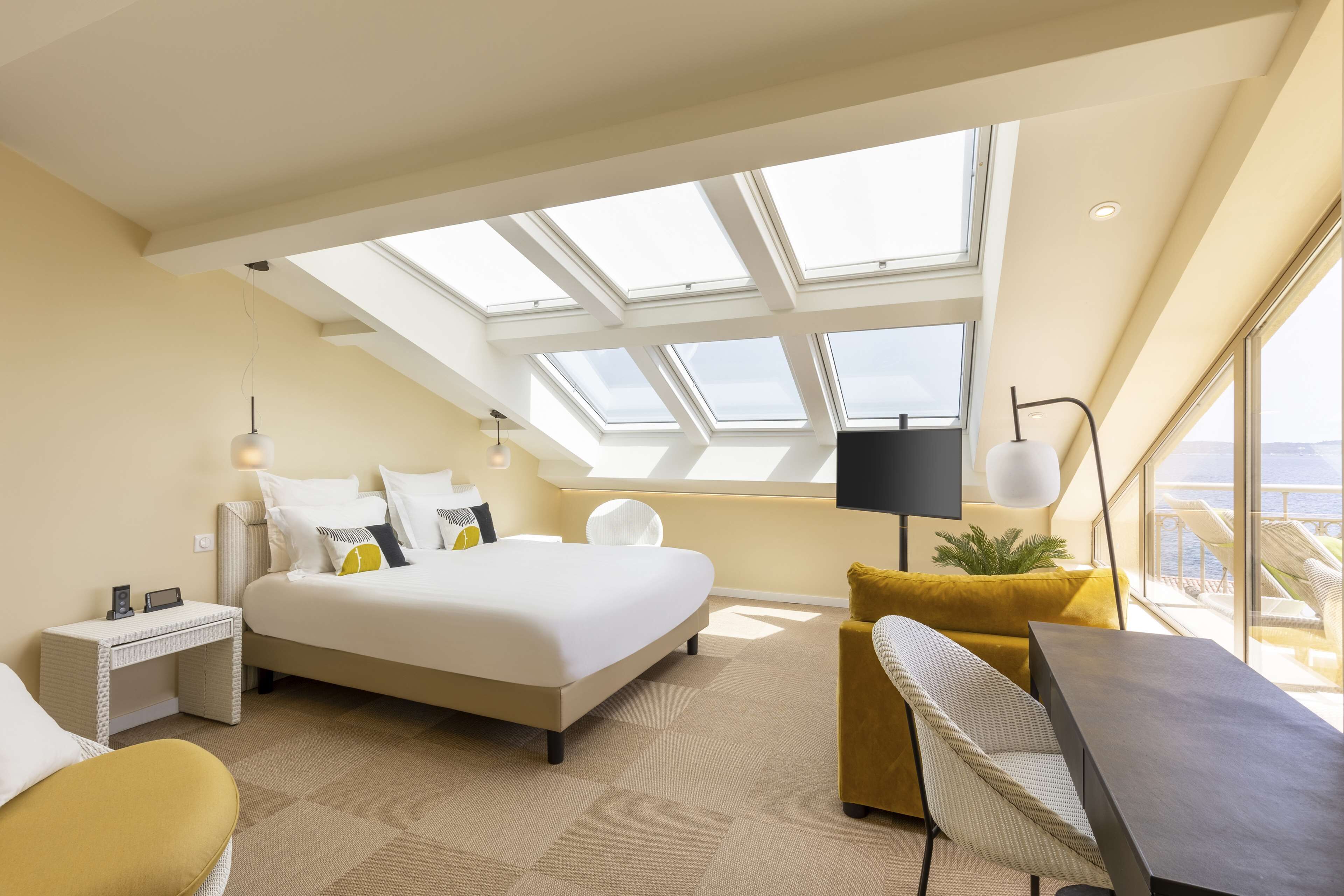 A modern hotel room with a skylight, a bed, a desk, a chair, and a view of the sea.