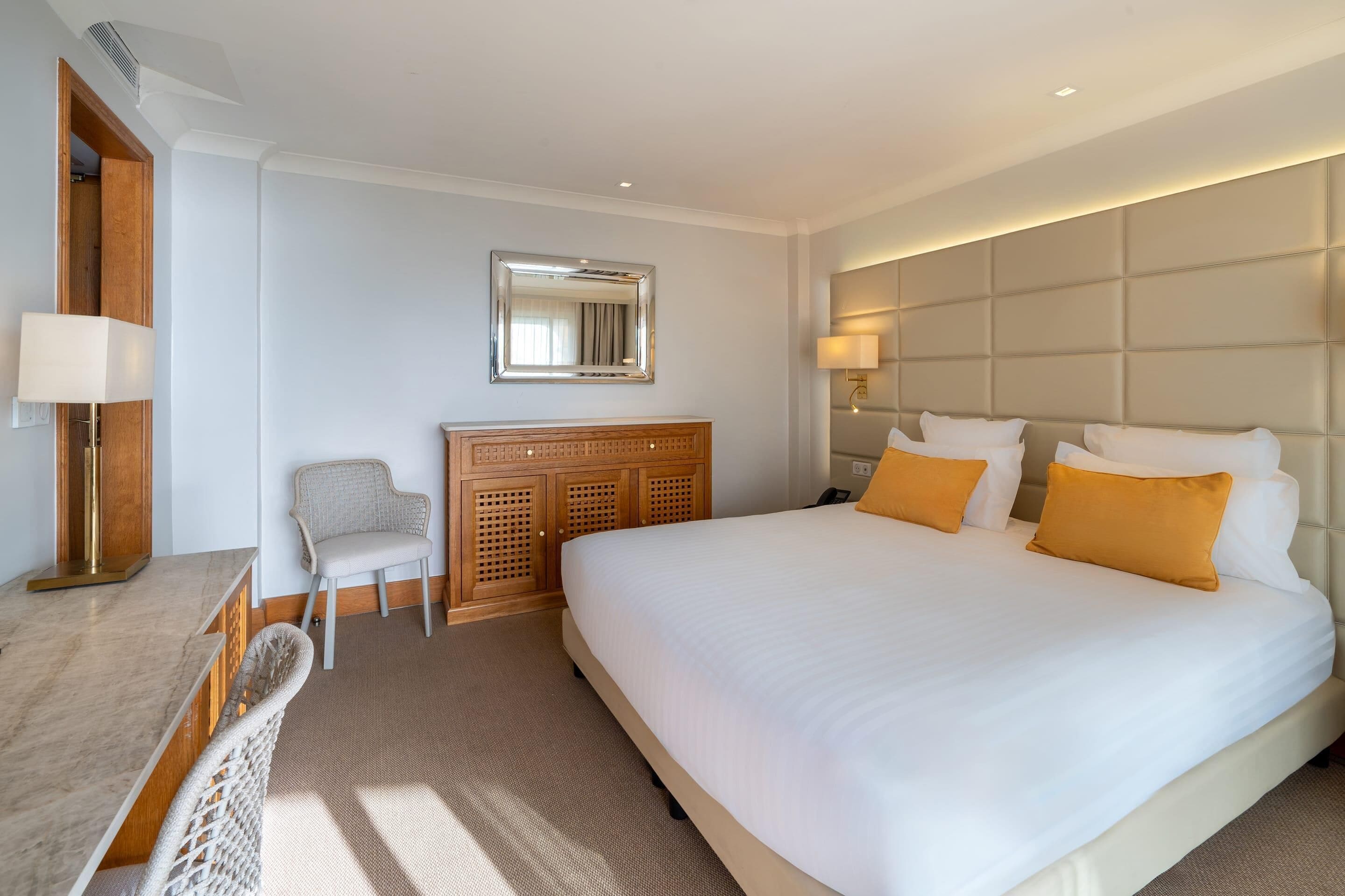 A hotel room with a large bed, a wooden headboard, a desk with a lamp, a chair, and a mirror on the wall.