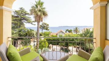 Een balkon met rieten meubilair en kussens, met uitzicht op een kustlandschap met bomen en de zee.