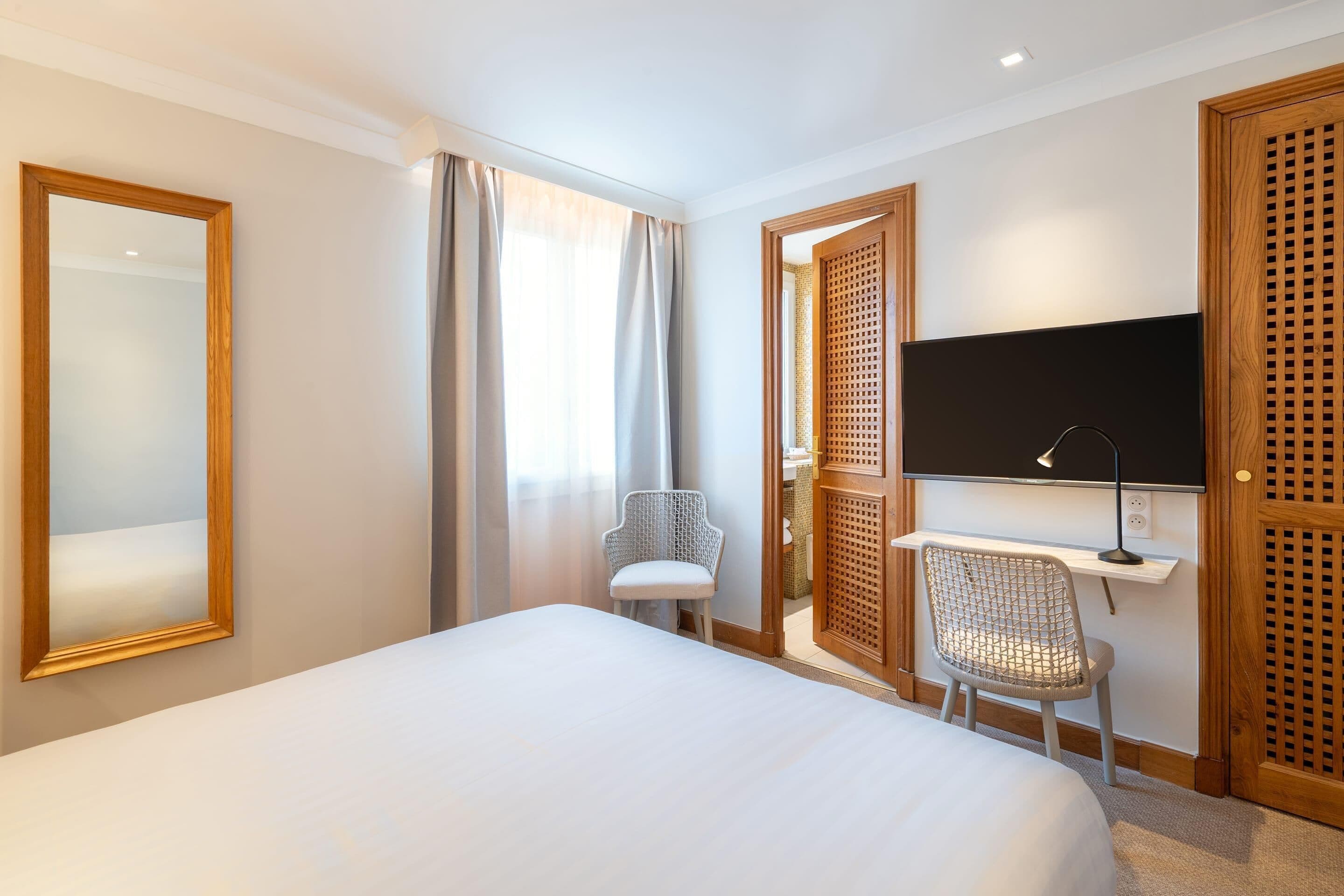A hotel room with a bed, a desk with a chair, a television, and a large mirror.