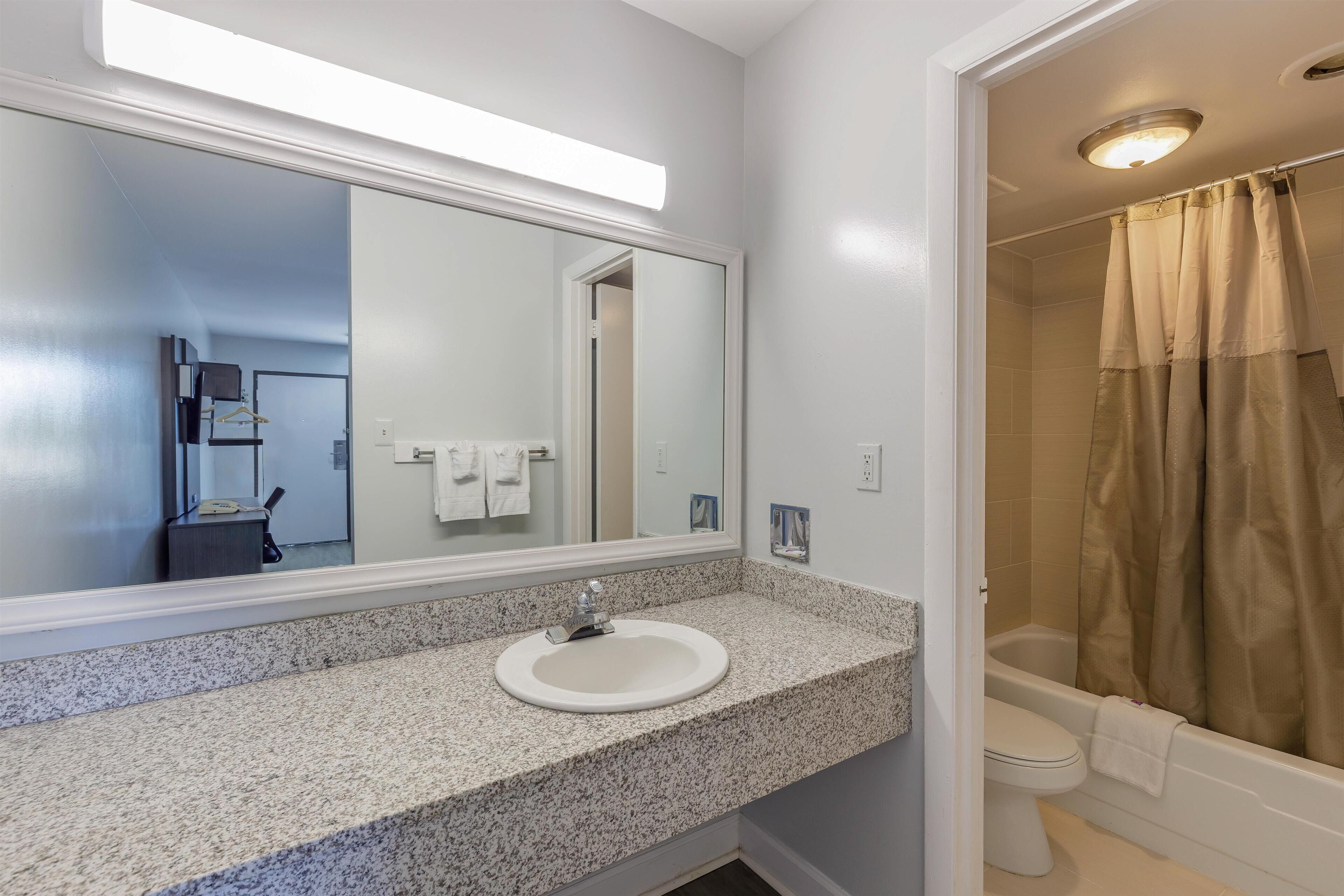 Une salle de bain avec un comptoir en granit, un lavabo blanc, un grand miroir et une douche avec des rideaux de couleur beige.