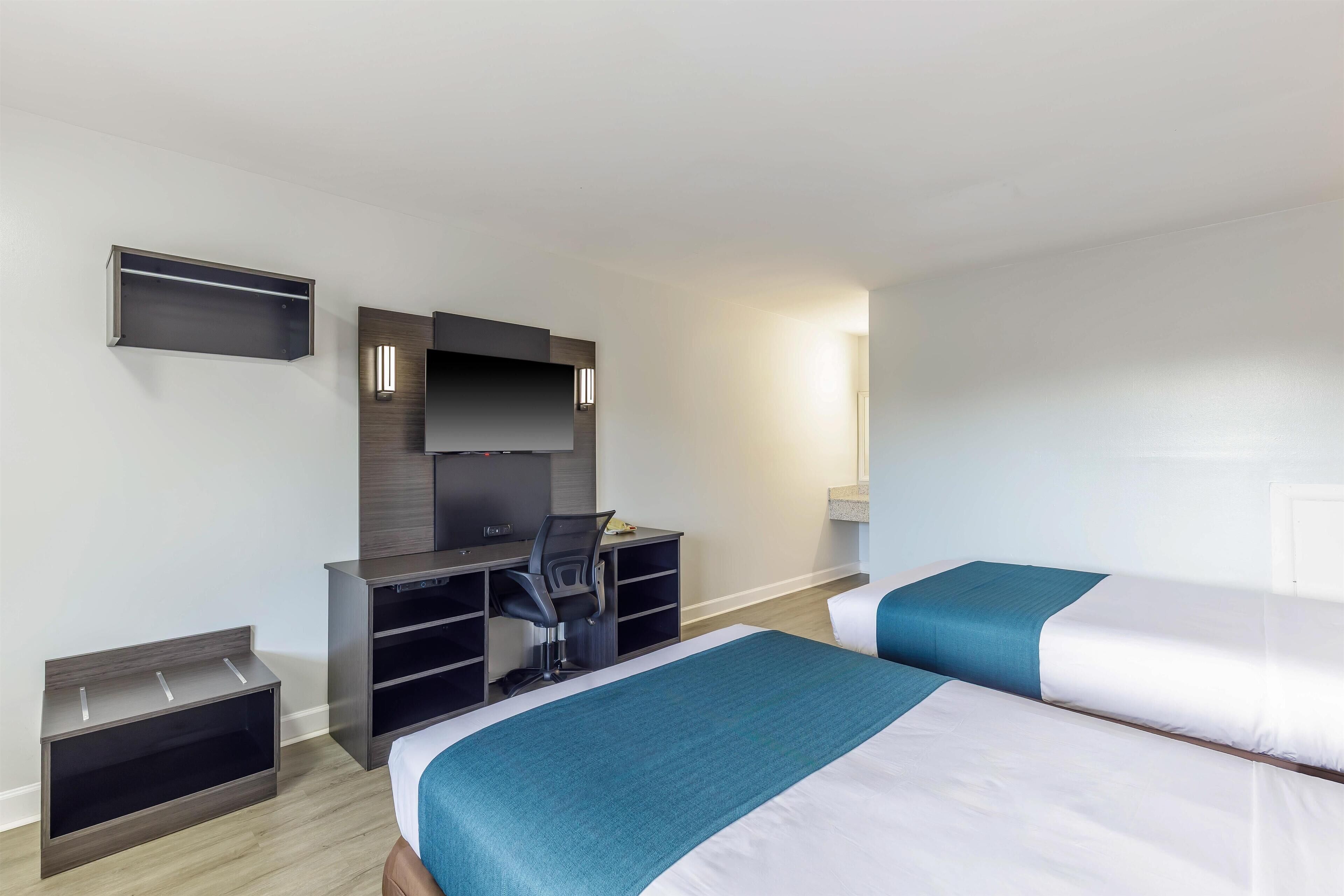 A hotel room with two beds, a desk with a chair, a TV, and a small shelf.