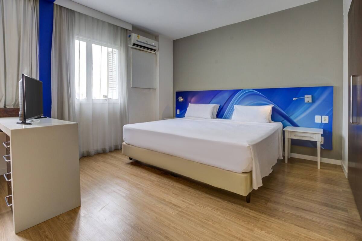 Habitación de hotel con cama, escritorio, silla, televisor y aire acondicionado instalado en la pared.
