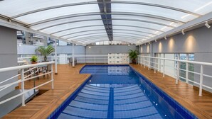 An indoor swimming pool with a glass ceiling, wooden deck, and potted plants.