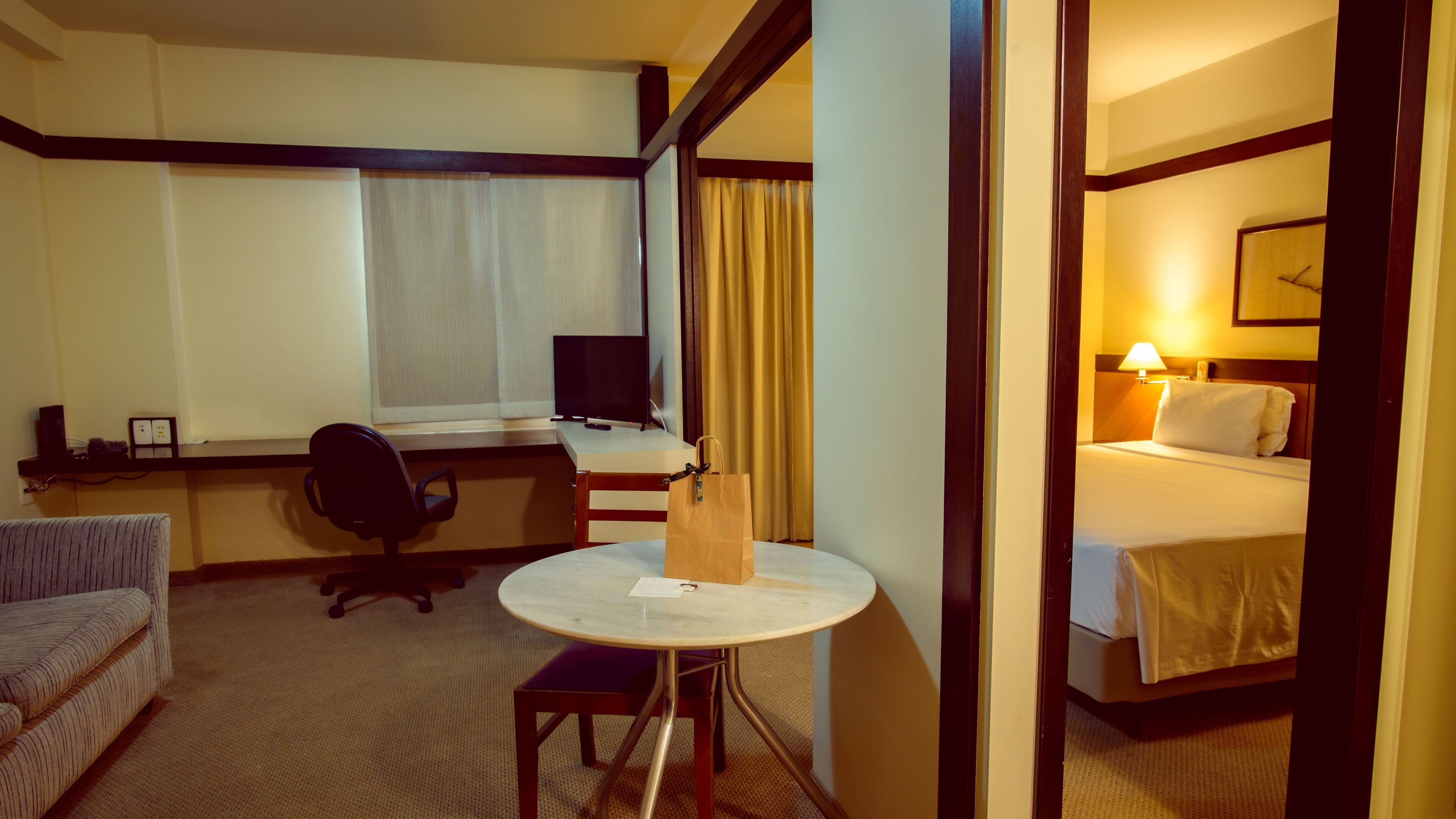 A hotel room with a bed, a desk with a computer, a chair, a small table, and a window with curtains.