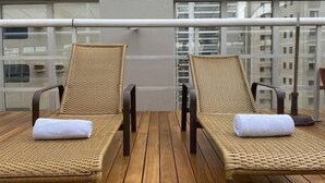 Two wicker lounge chairs with towels on a wooden deck, a glass railing, and a building in the background.