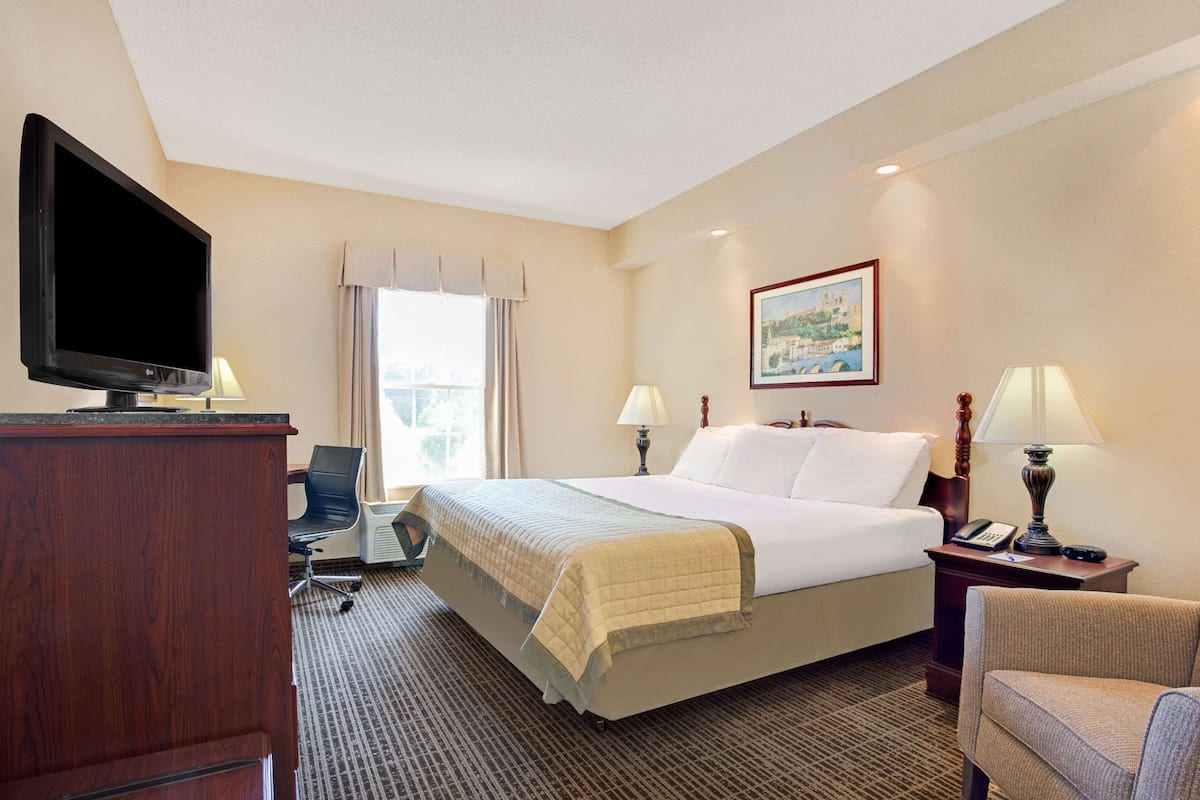 A hotel room with a bed, a desk with a television, two lamps, a chair, and a painting on the wall.