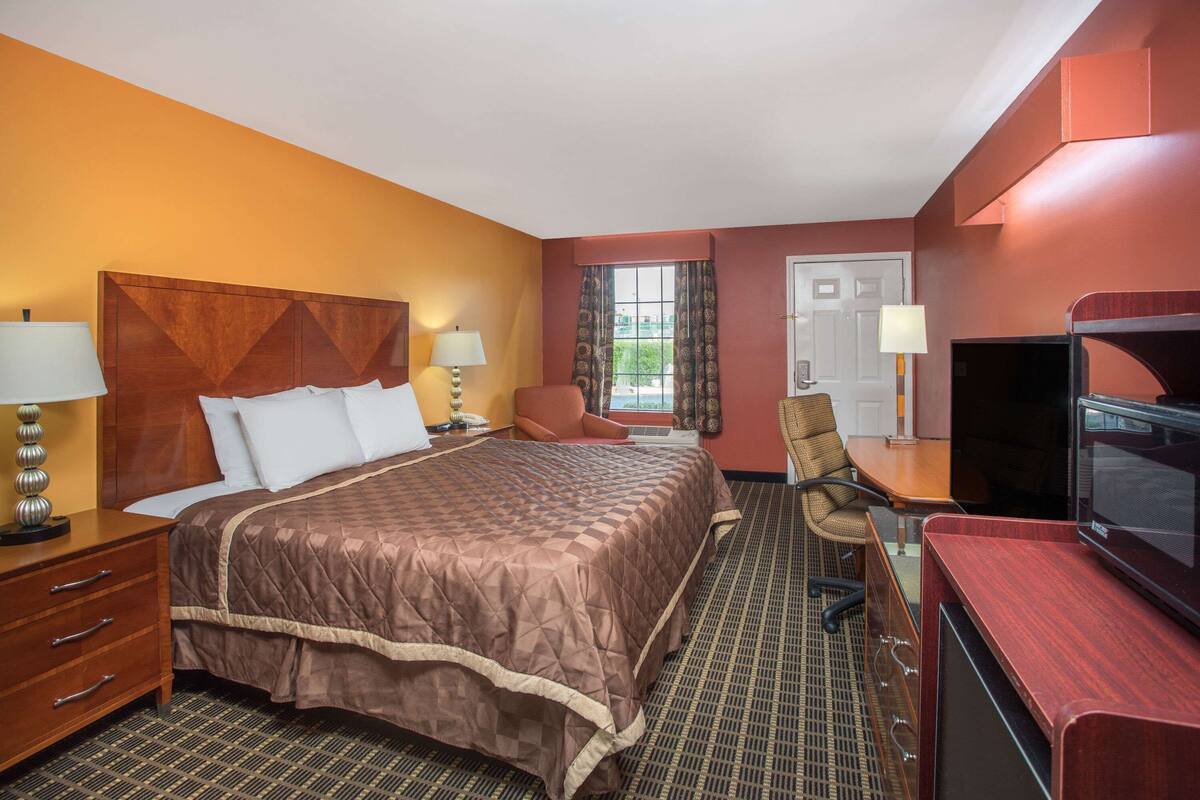 Ein Hotelzimmer mit einem Bett, zwei Stühlen, einem Schreibtisch mit Fernseher, einem Schminktisch und einem Fenster mit Vorhängen.
