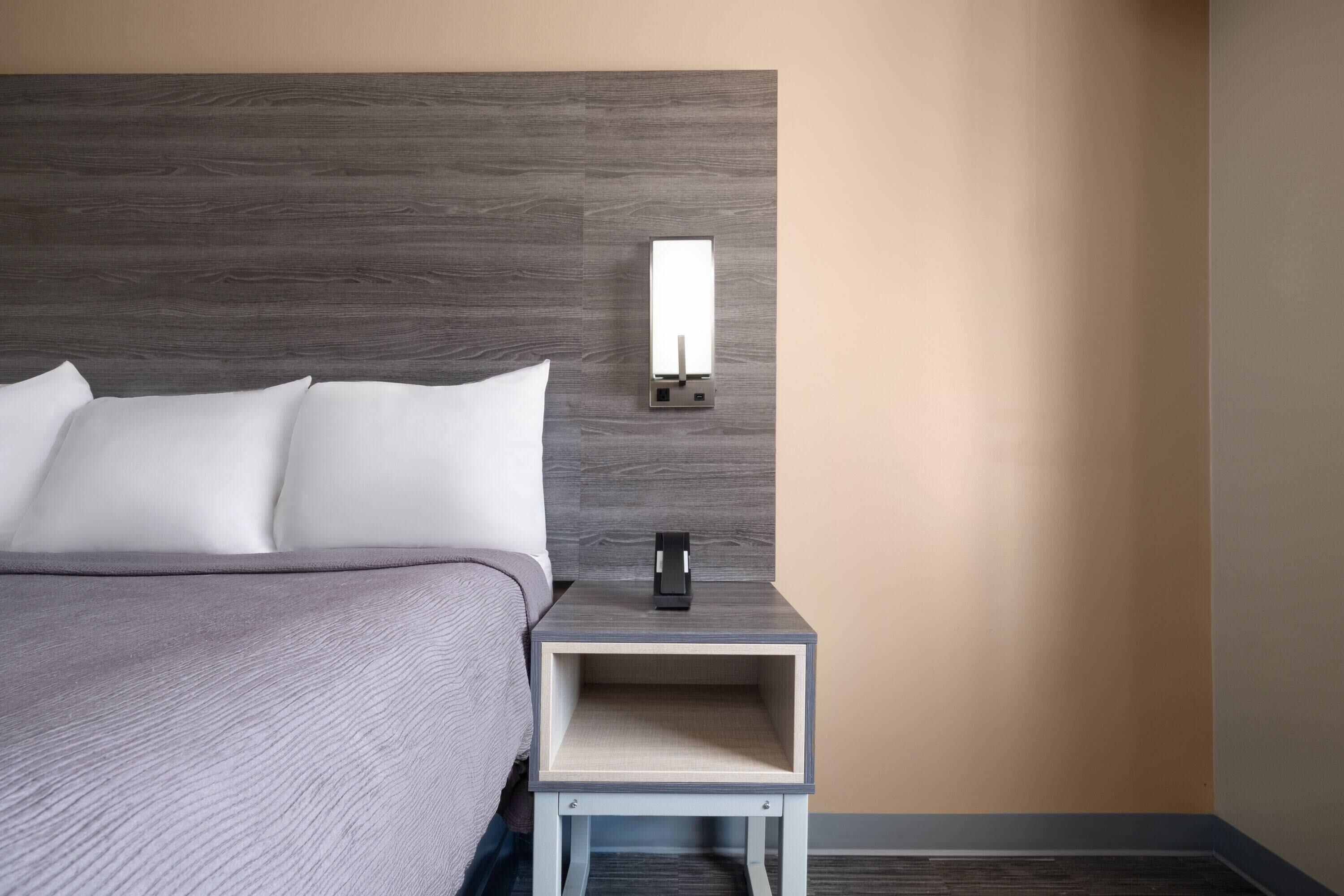 Una cama bien hecha con sábanas blancas, una mesita de noche con lámpara, y un aplique en la pared.